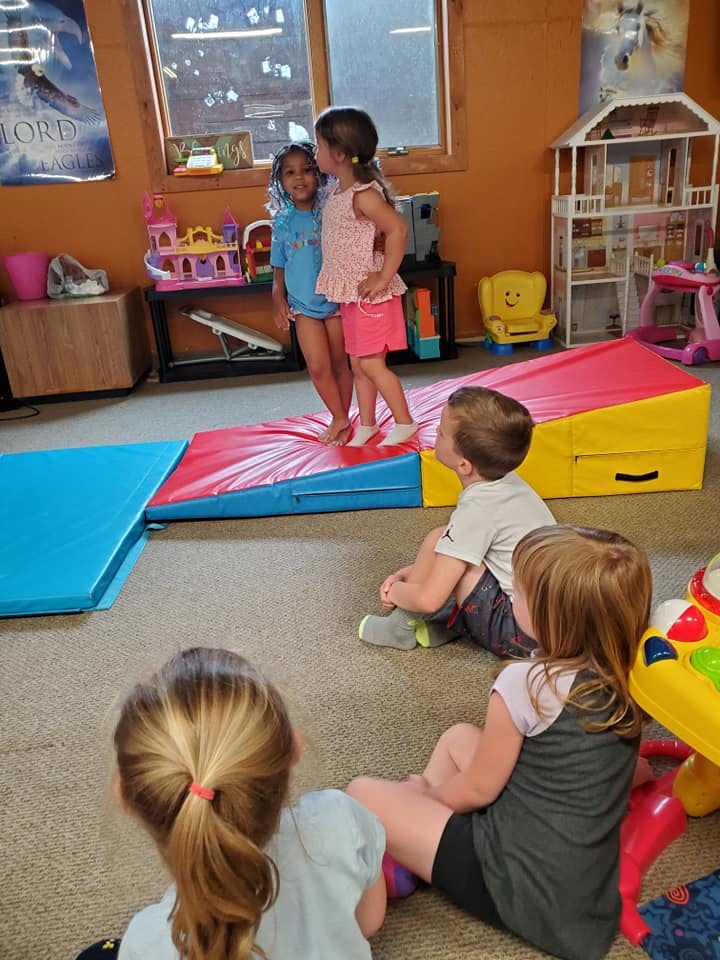 Two children standing on a soft play mat, other children sitting on the floor
