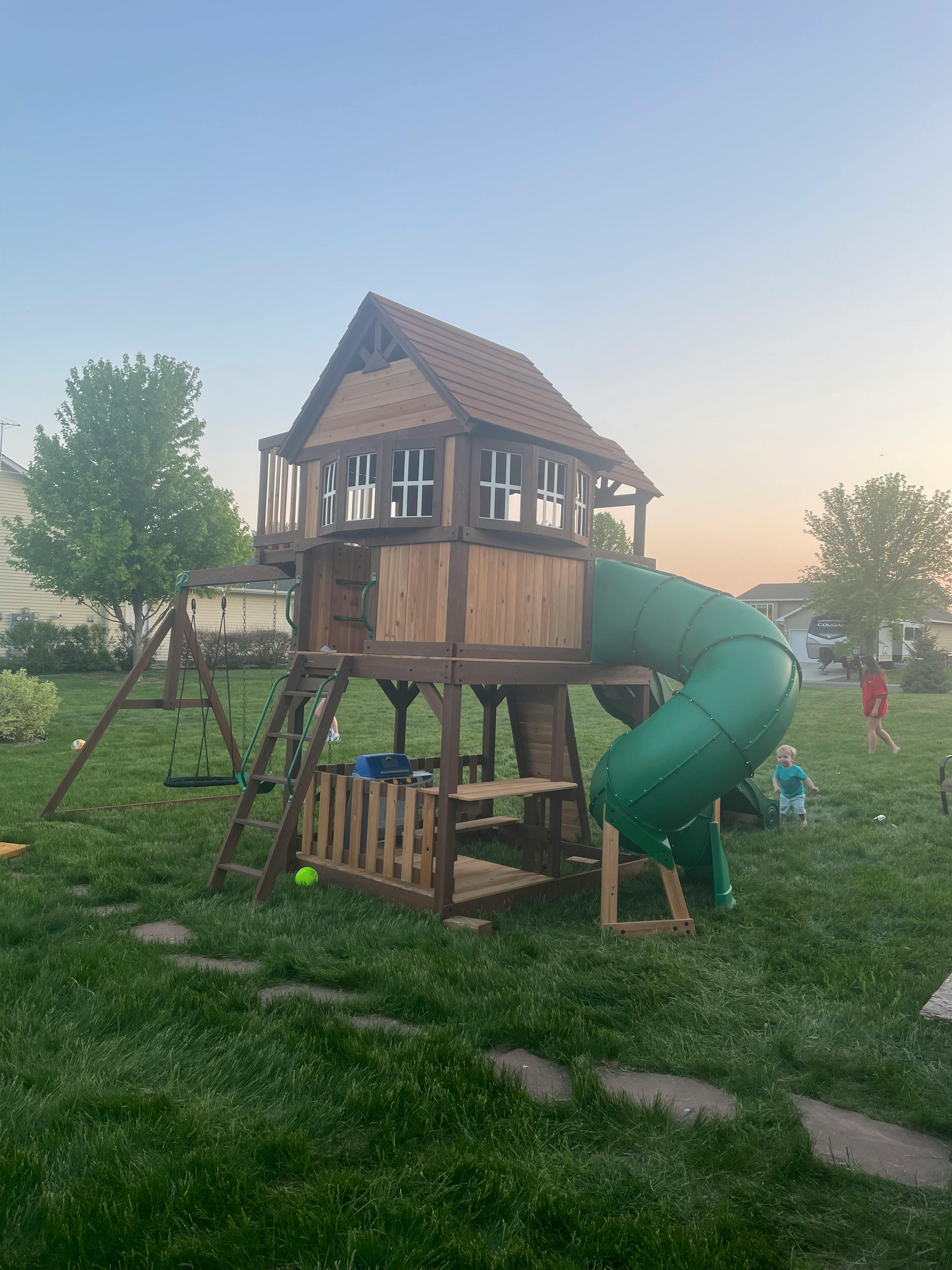 Wooden playset with a house, slide, and swings in a grassy yard