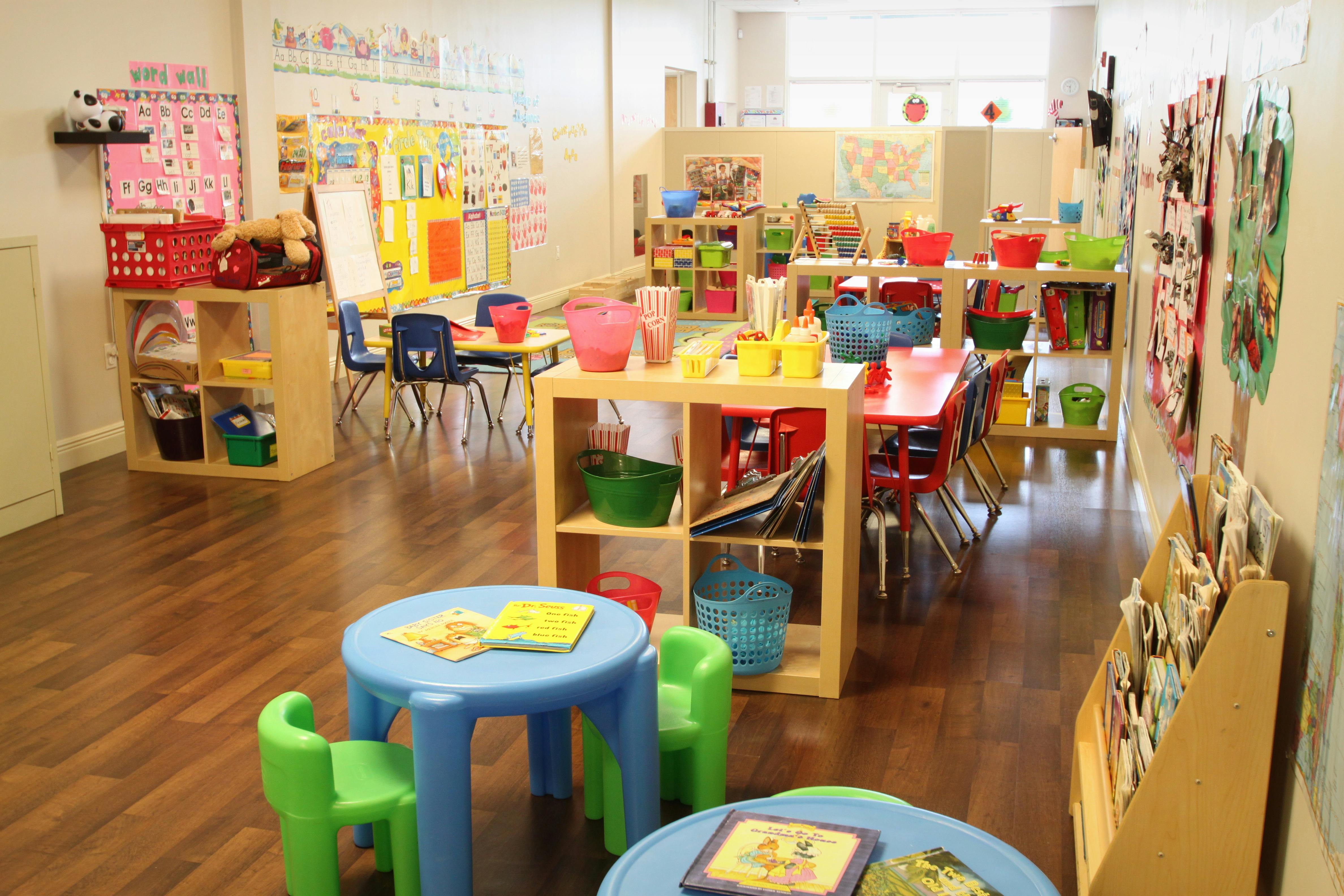 Classroom with small tables and chairs, shelves with toys and art supplies