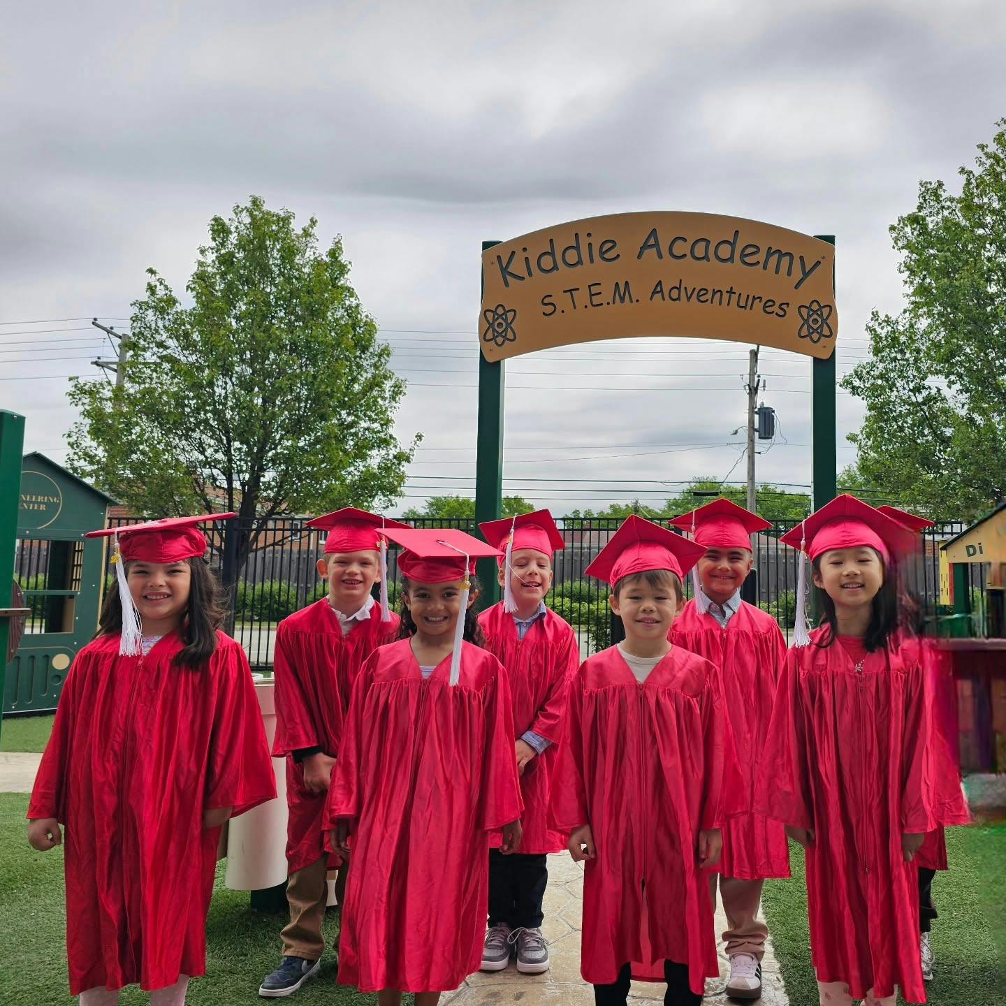 Group of children wearing red graduation caps and gowns by a Kiddie Academy sign