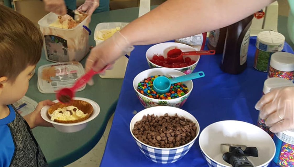 Person serving ice cream to a child with toppings on a table