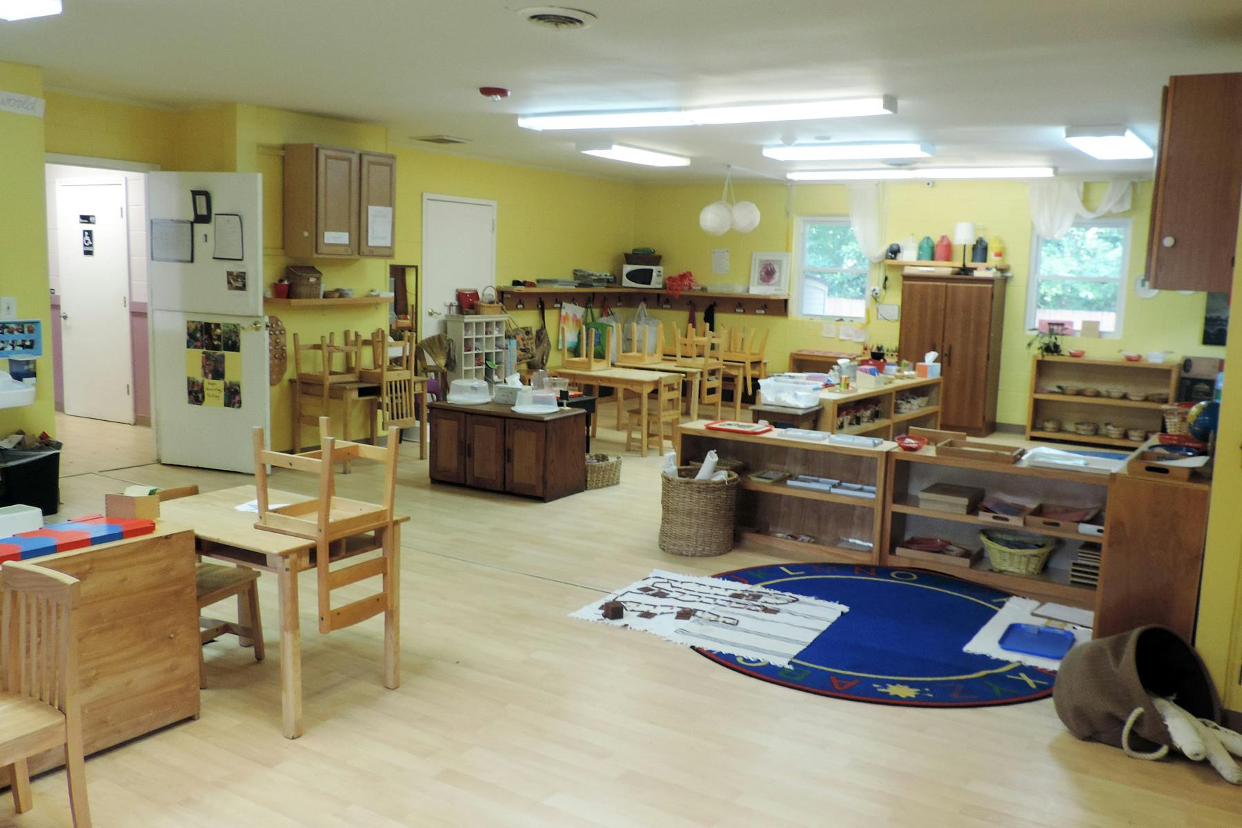 Classroom with small wooden tables and chairs, shelves with learning materials