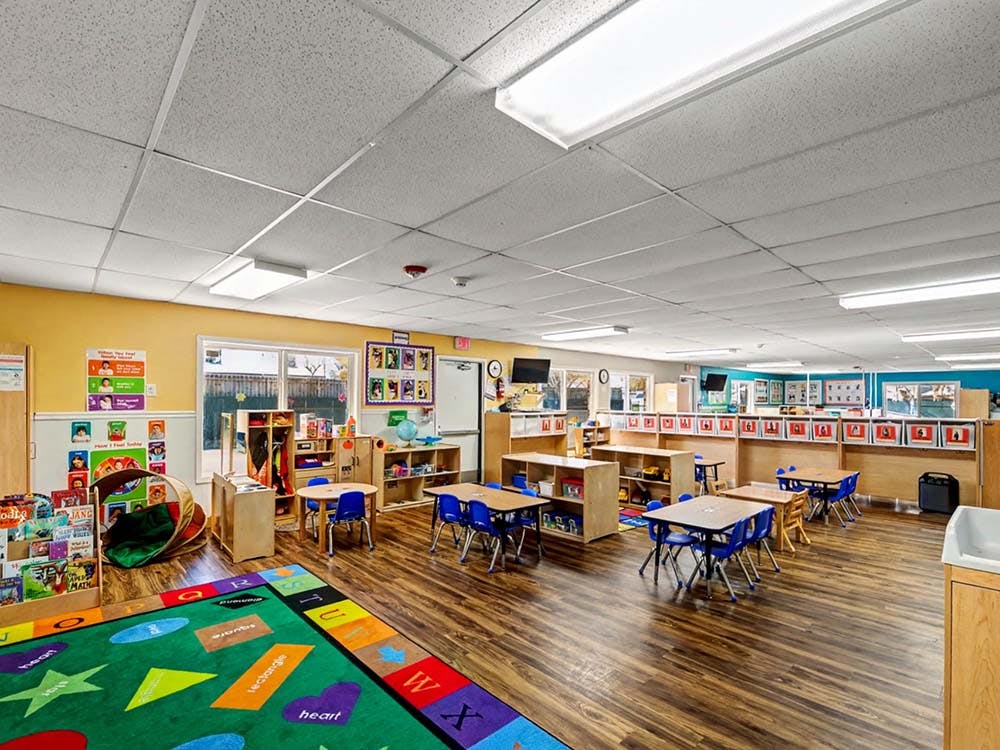 Empty classroom with small tables and chairs, a colorful rug, and windows