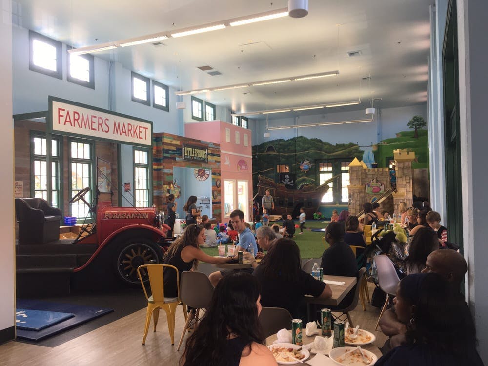 People eating at tables in a room with a "Farmers Market" sign and play structures