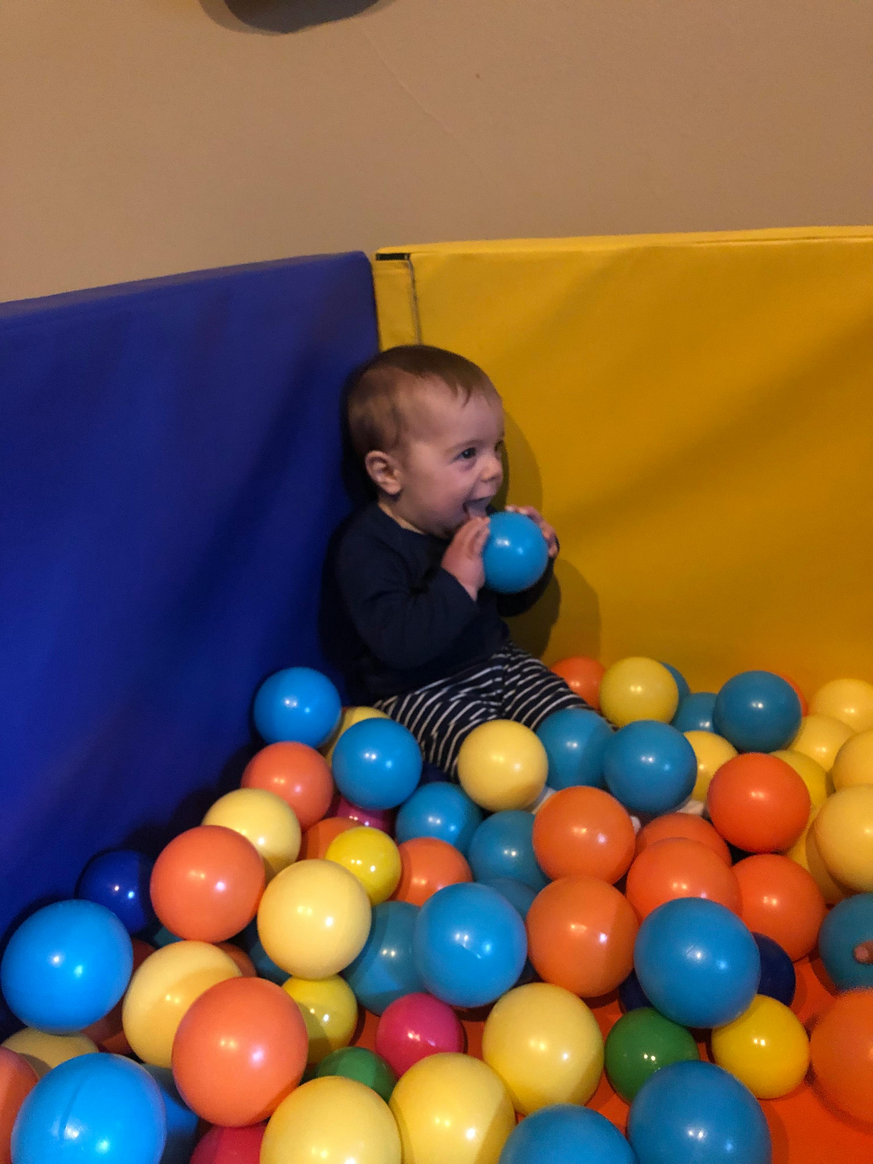 Child sitting in a ball pit, holding a blue ball to their mouth
