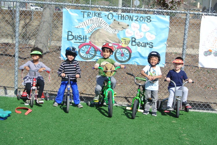 Five children on tricycles and bicycles in front of a banner that reads "TRIKE-O-THON 2018"