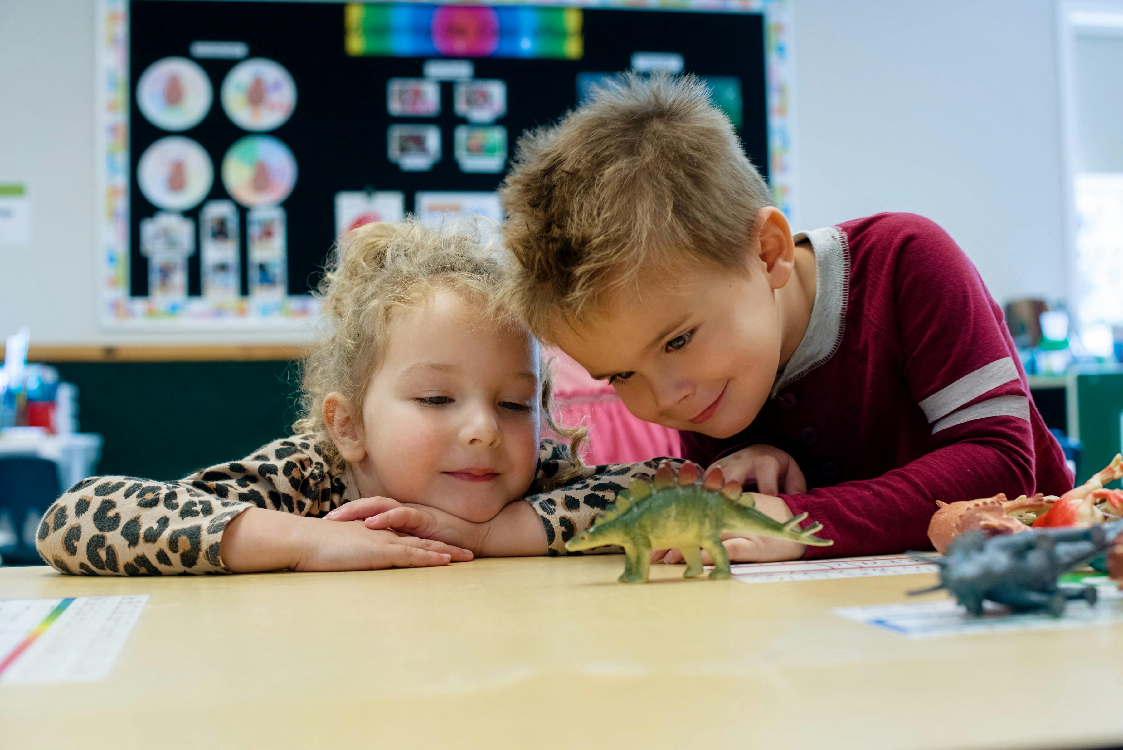 Two children looking at a toy dinosaur on a table in a classroom