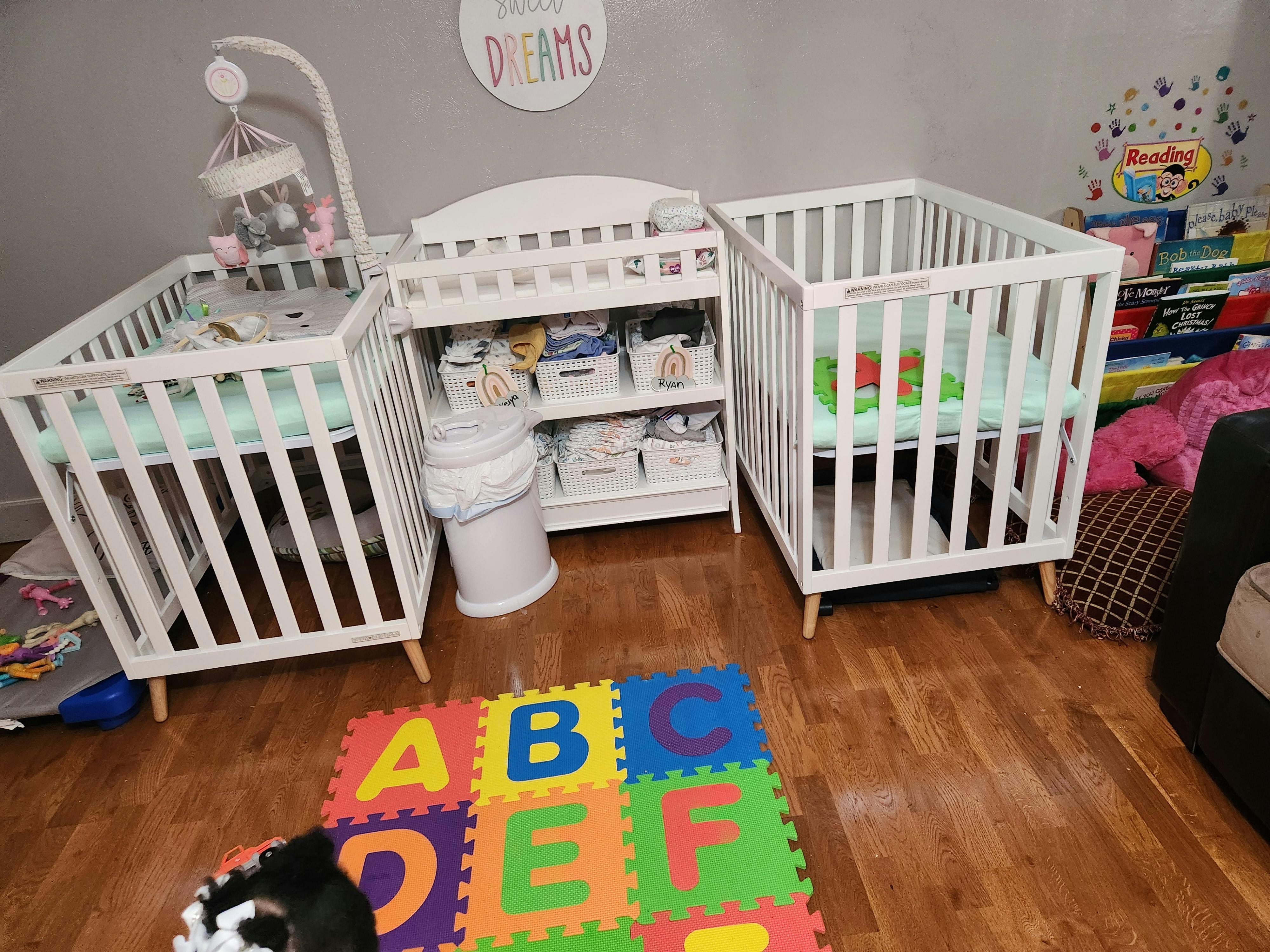 Two white cribs with a changing table in a room with an alphabet playmat