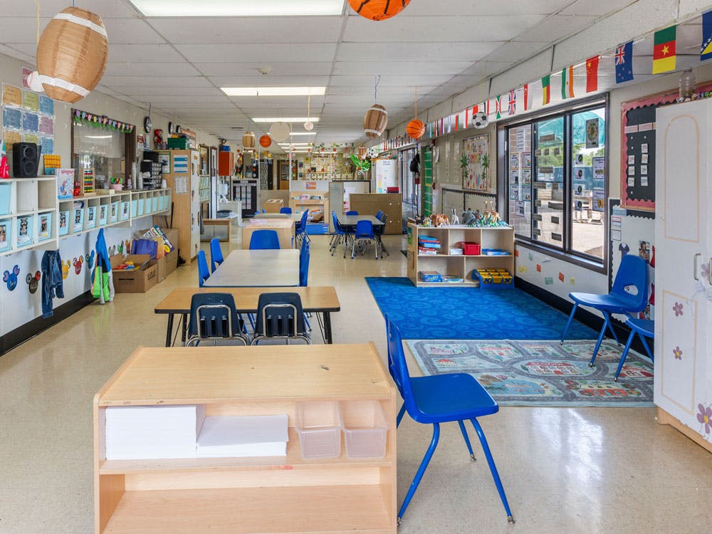 Classroom with small tables and chairs, shelves, and a window with flags above