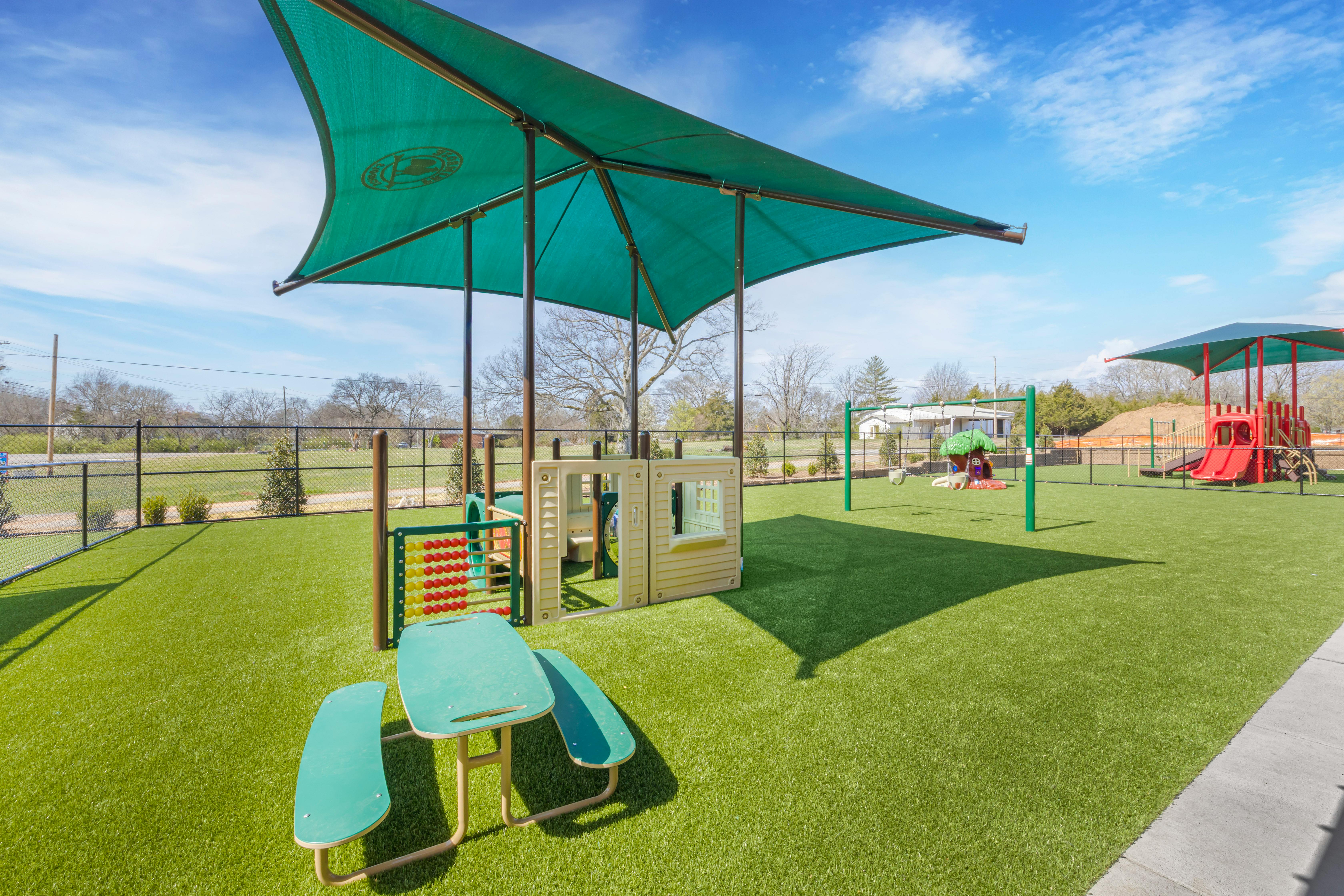 Playground with green turf, a climbing structure, swings, and shade covering over a picnic table