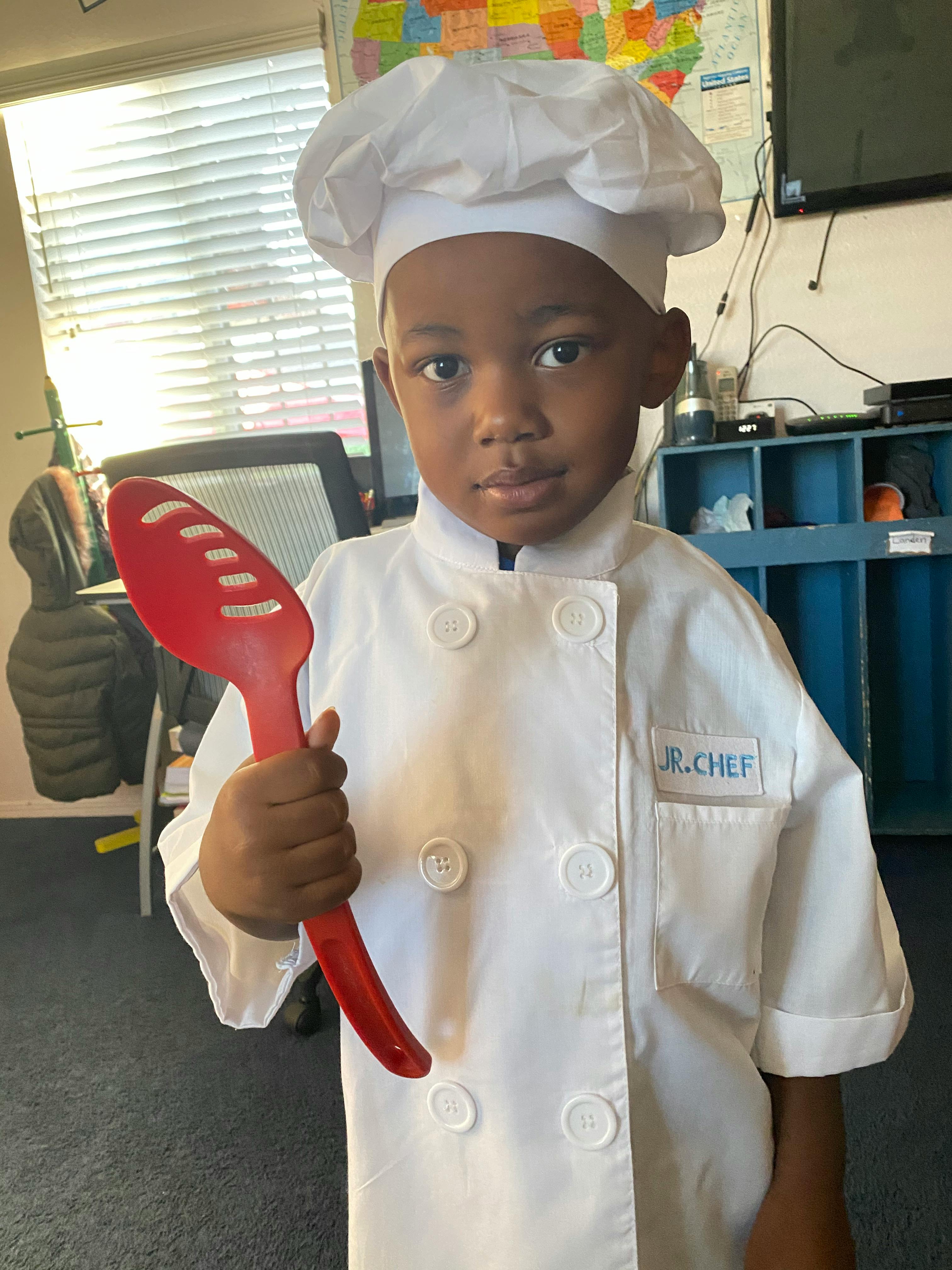 Child wearing a chef uniform and hat holding a red plastic cooking spoon