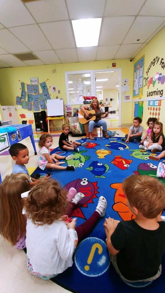 Adult playing guitar for children sitting on a fish-patterned rug in a classroom