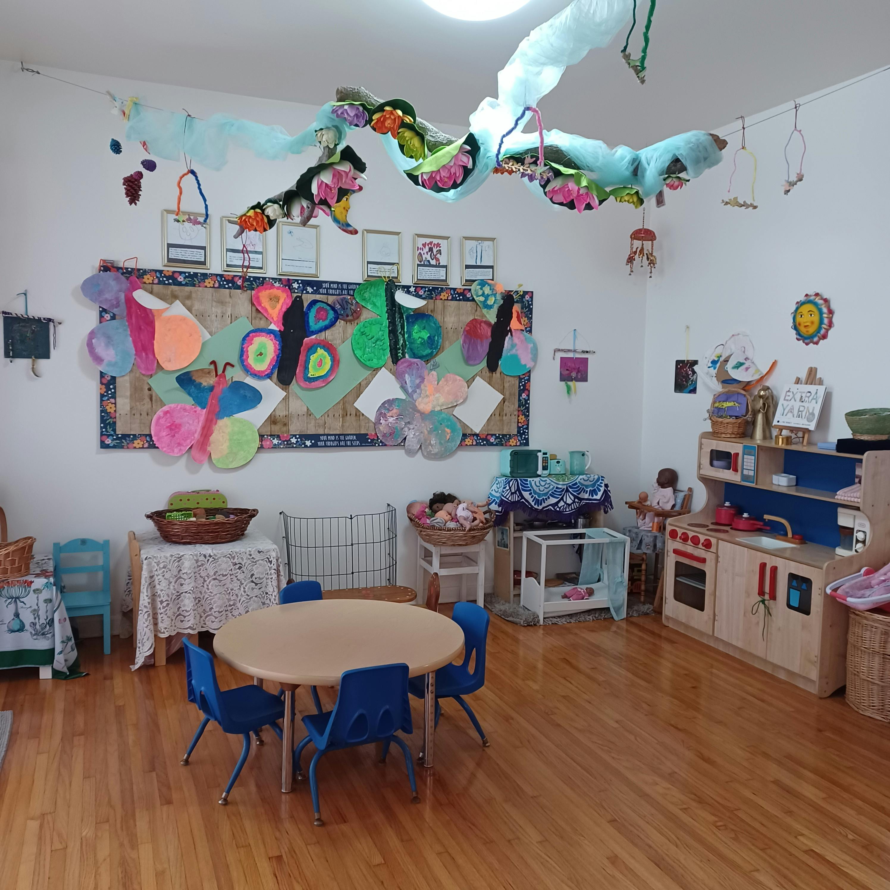 Classroom with a small table and chairs, play kitchen, and bulletin board with crafts