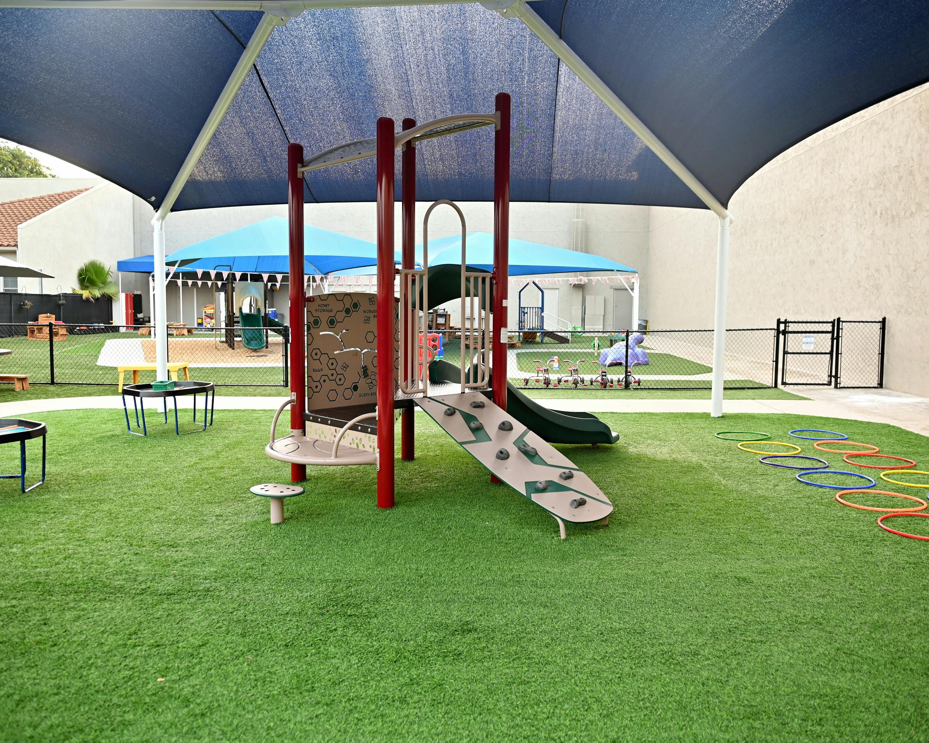 Outdoor playground with artificial turf, covered by a large blue shade structure