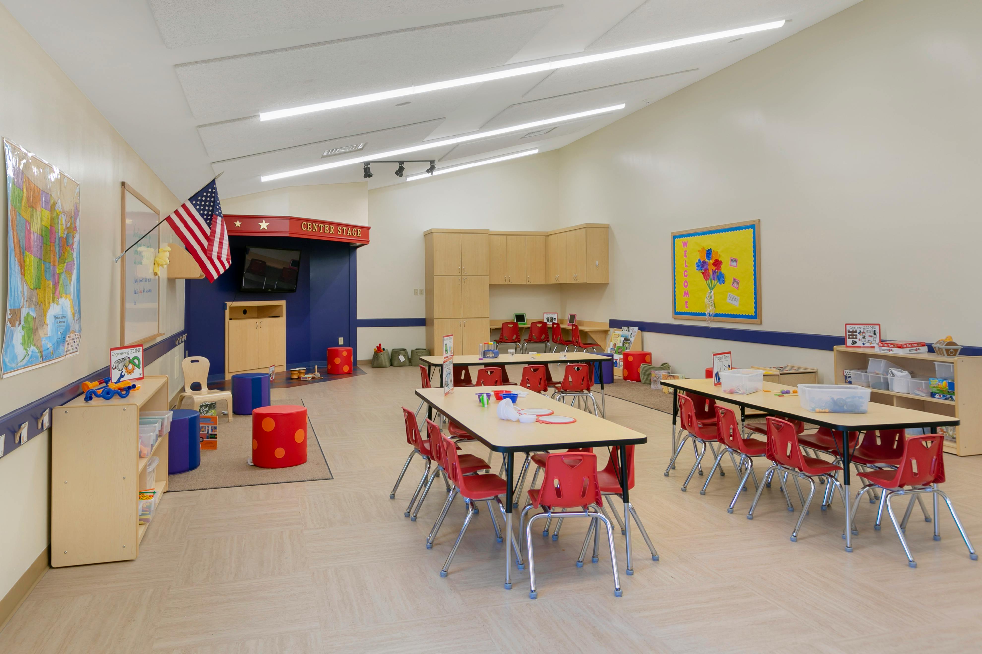 Classroom with small tables and red chairs, an American flag, and a small stage