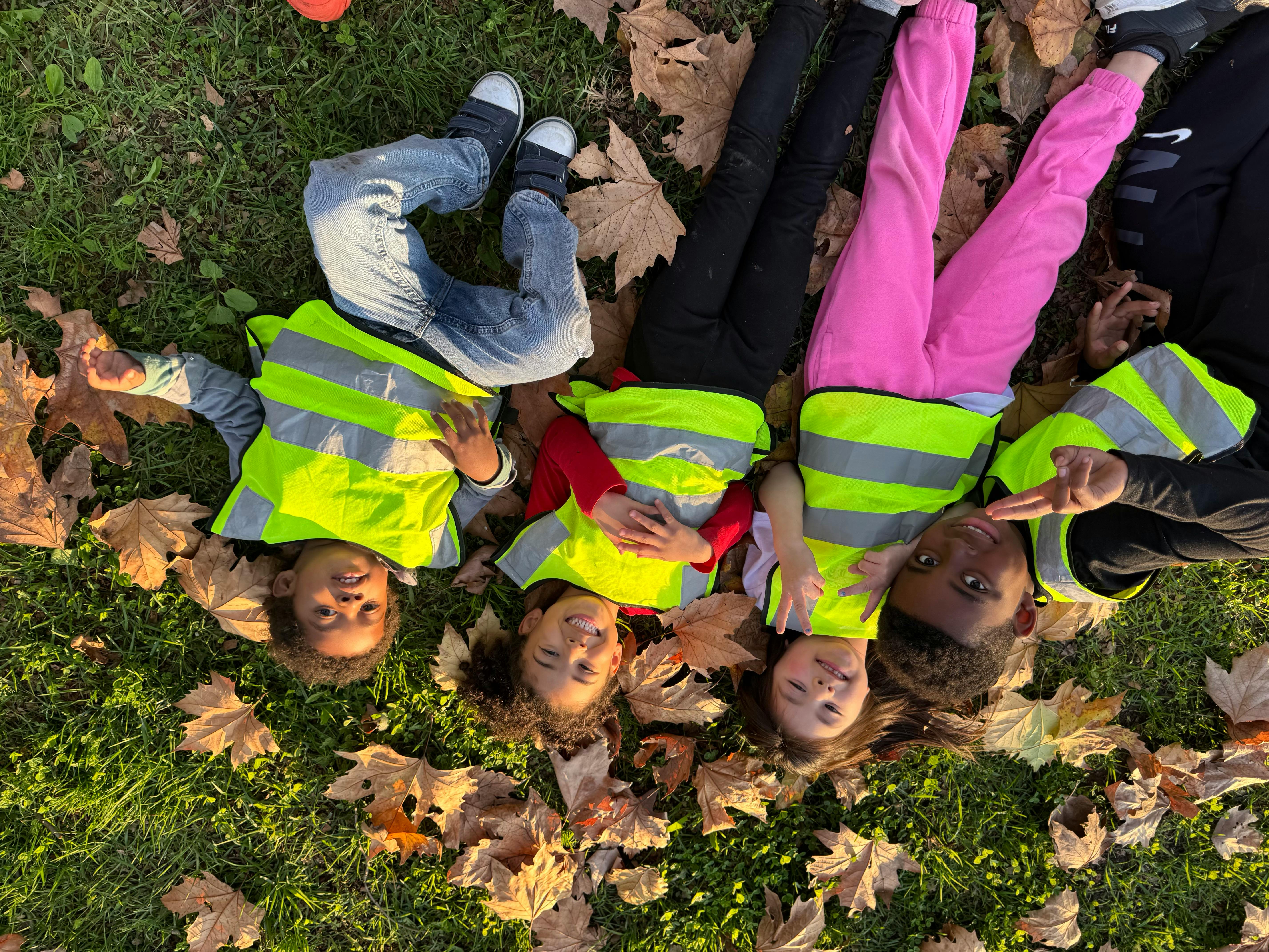 Four children in safety vests lying on grass with autumn leaves