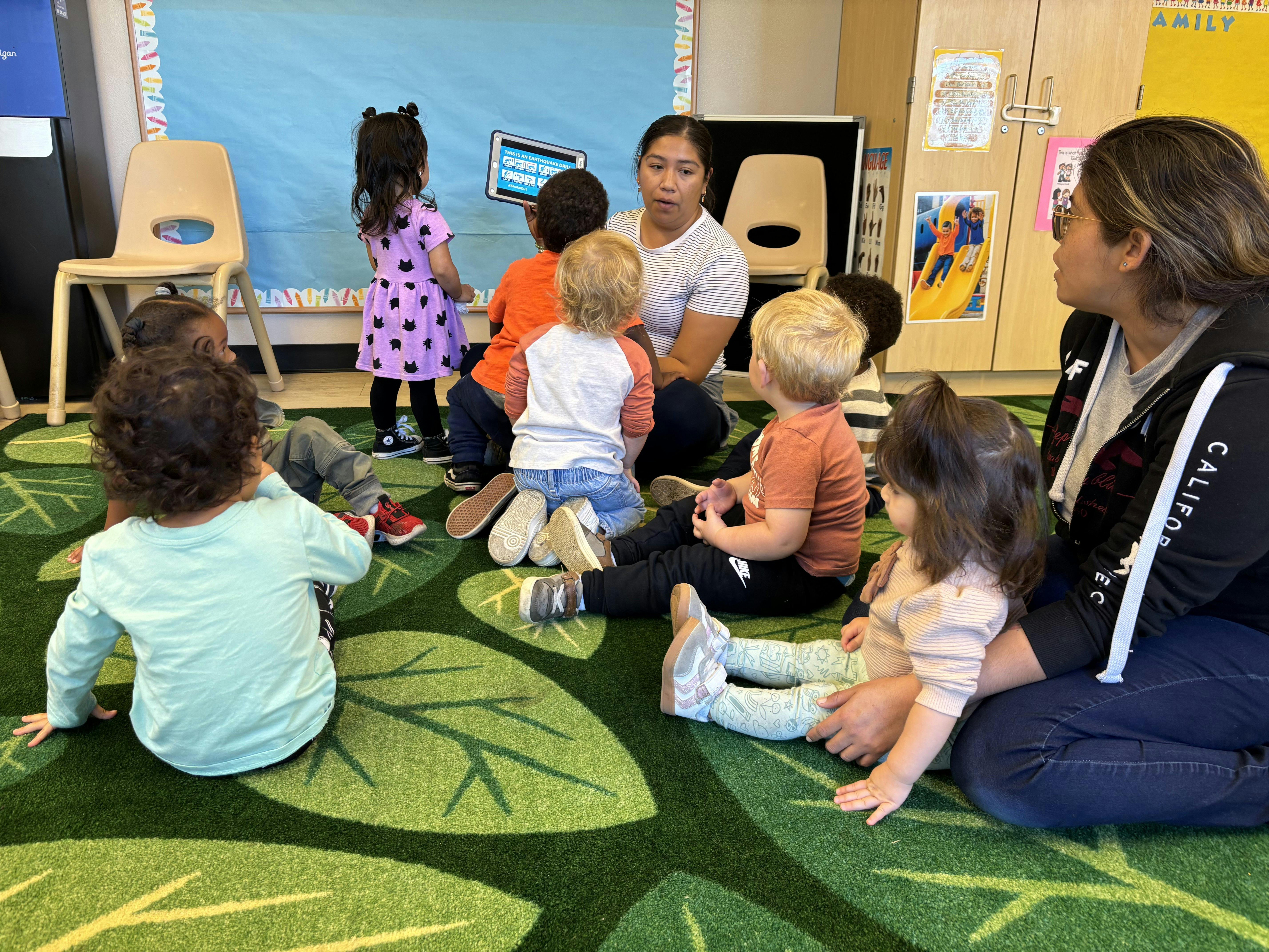 Adult and several children sitting on a leafy green rug, looking at a tablet