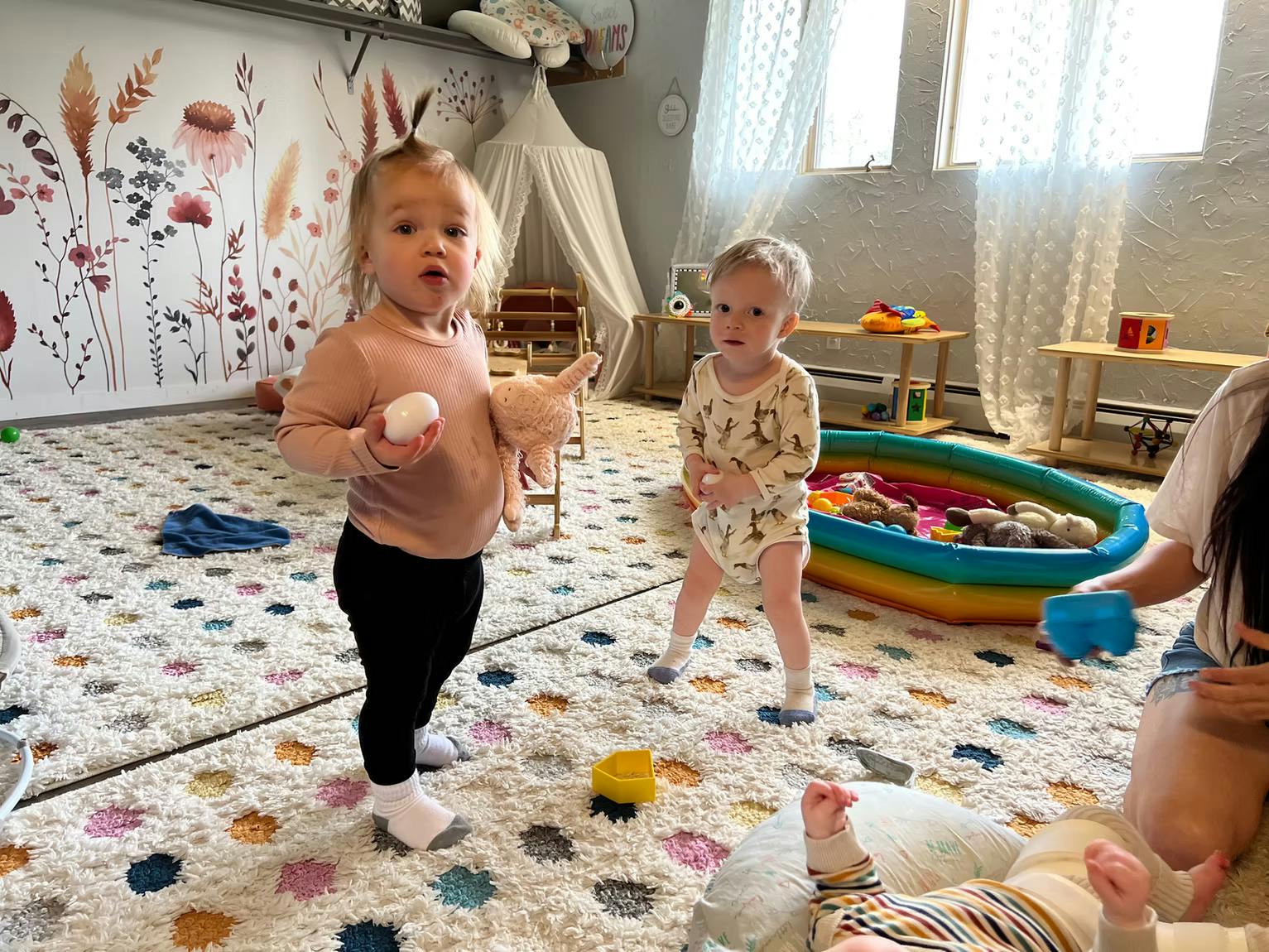 Two children standing in a playroom with toys and a person sitting on the floor