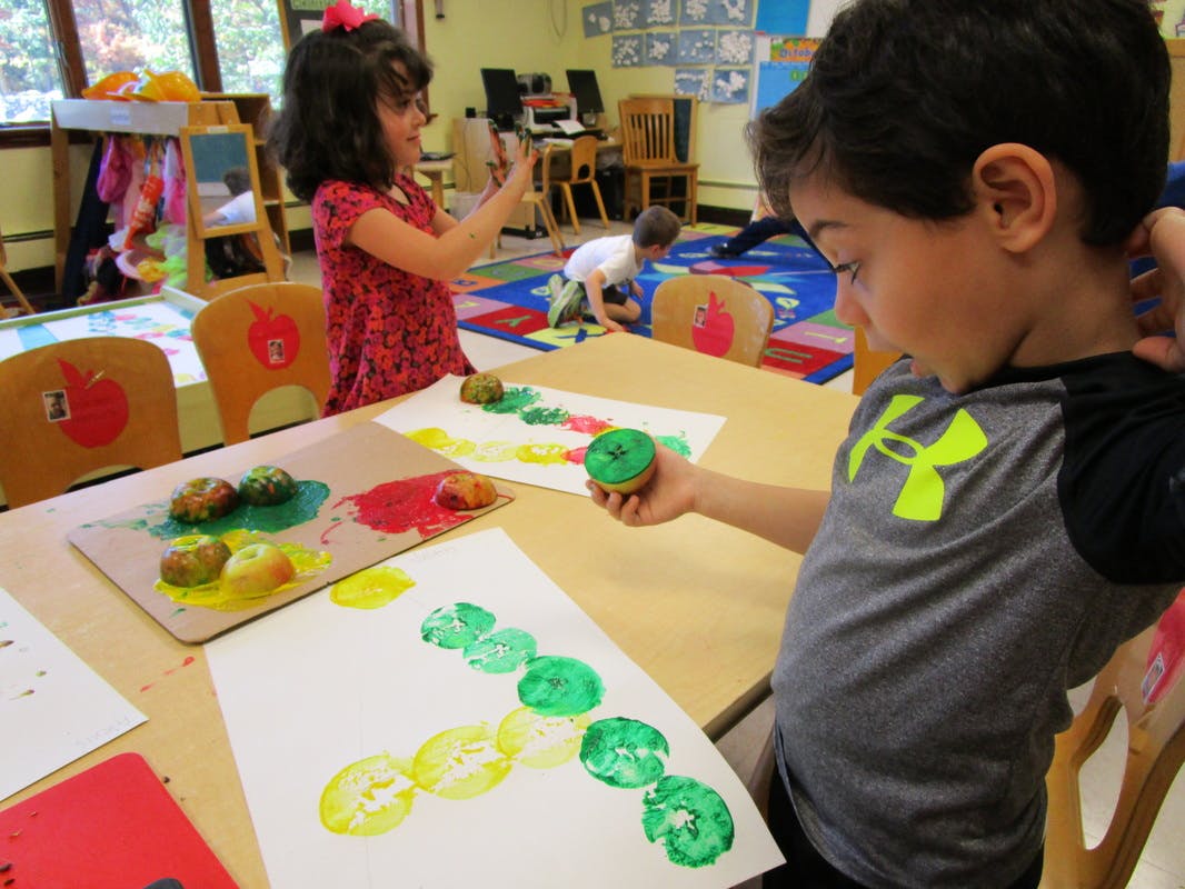 Child holding a painted apple, another child painting in a classroom