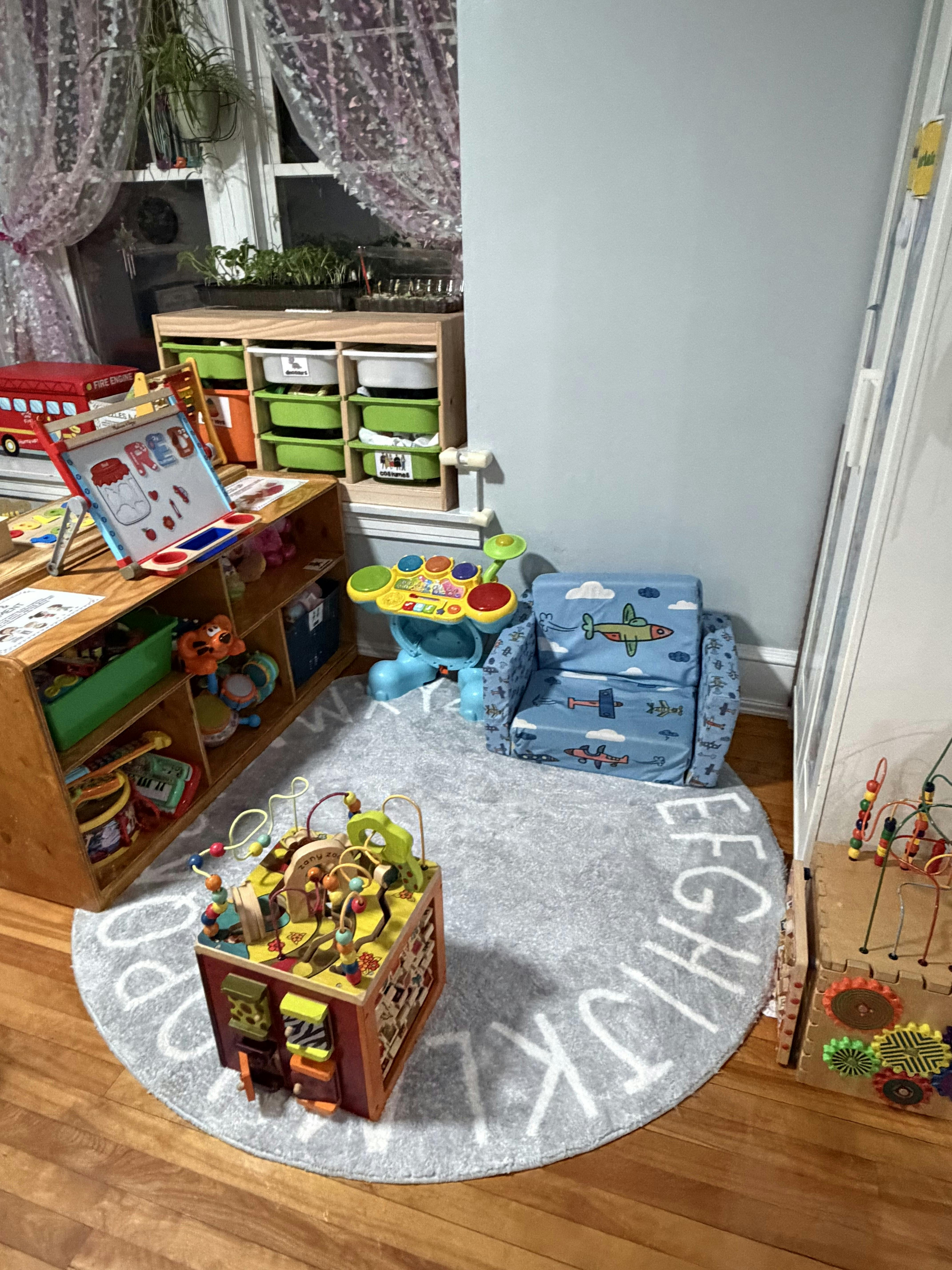 A playroom with various colorful toys, storage shelves, and a gray rug with letters