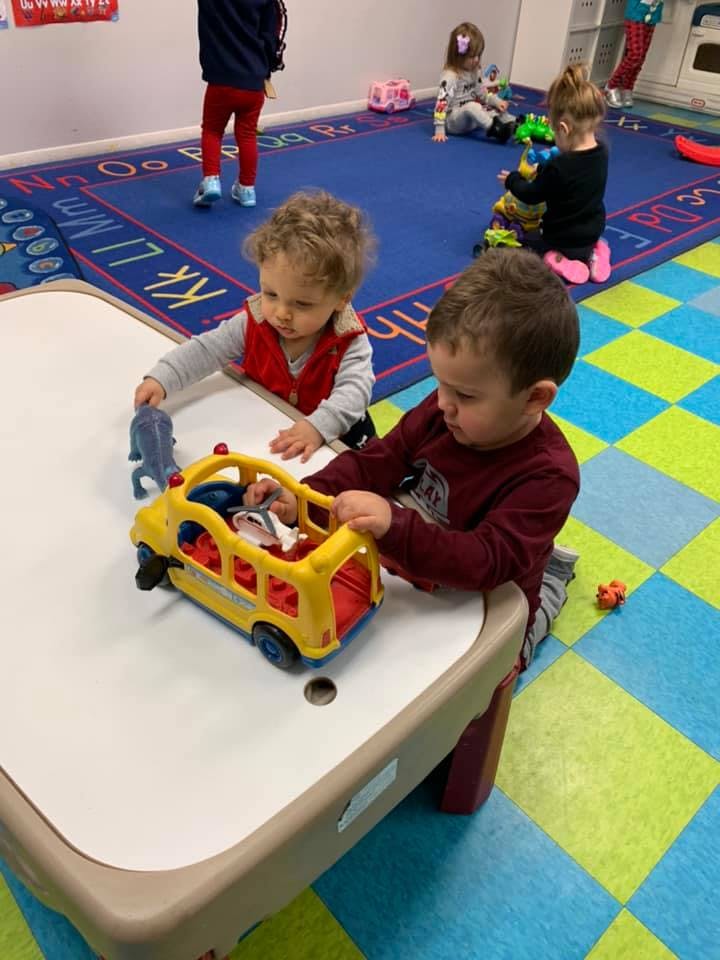 Two children playing with a toy bus at a table in a classroom