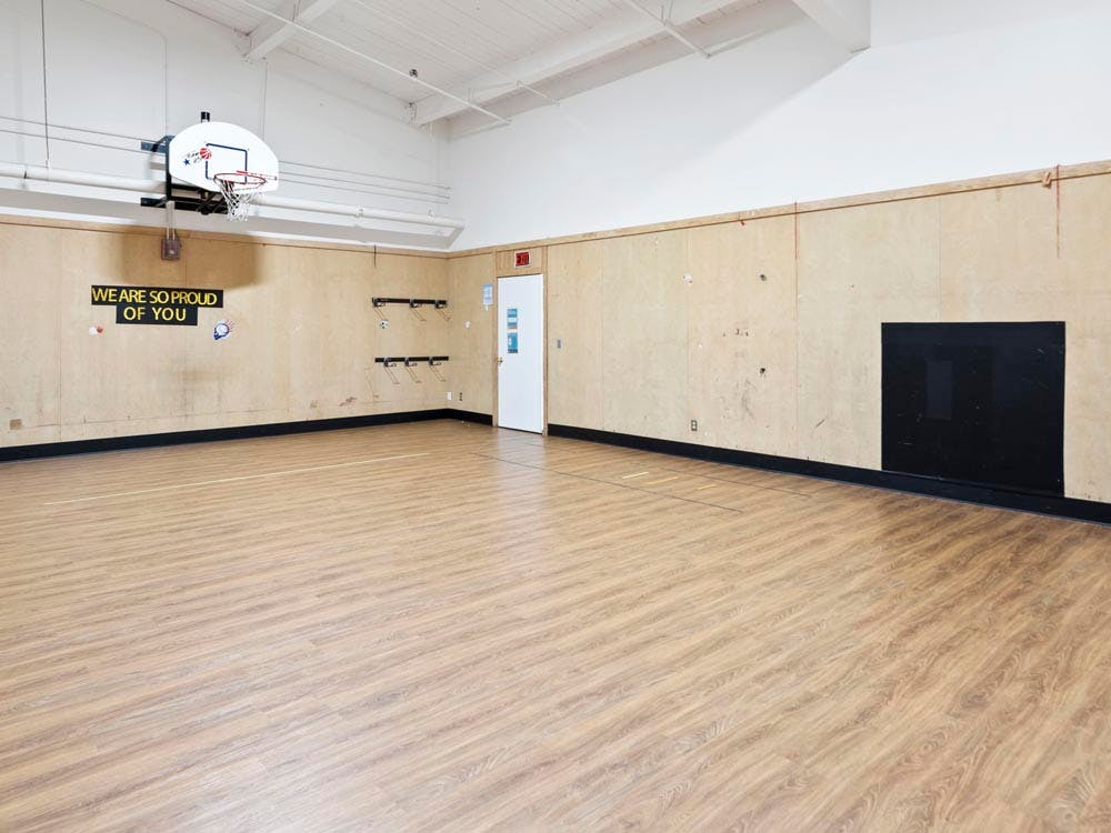 Empty gymnasium with a basketball hoop, wooden walls, and sign "WE ARE SO PROUD OF YOU"