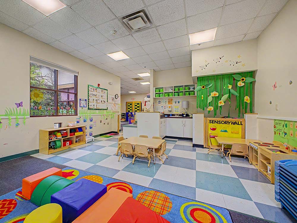 A classroom with small tables and chairs, a sensory wall, and play mats