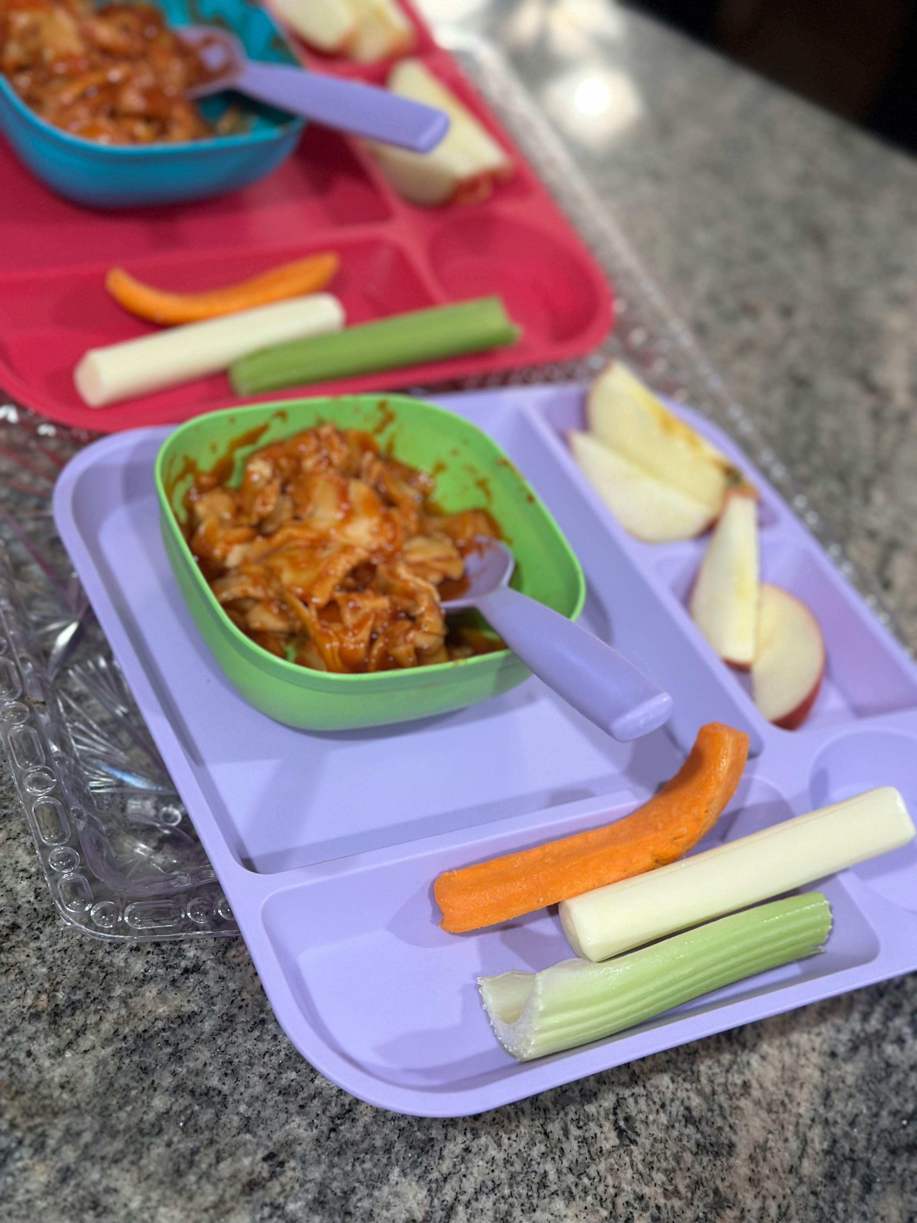Two divided plates with food, including a main dish, apple slices, and vegetable sticks