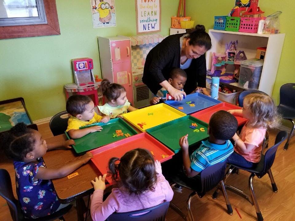 Adult supervising children playing with colorful trays at a table in a classroom