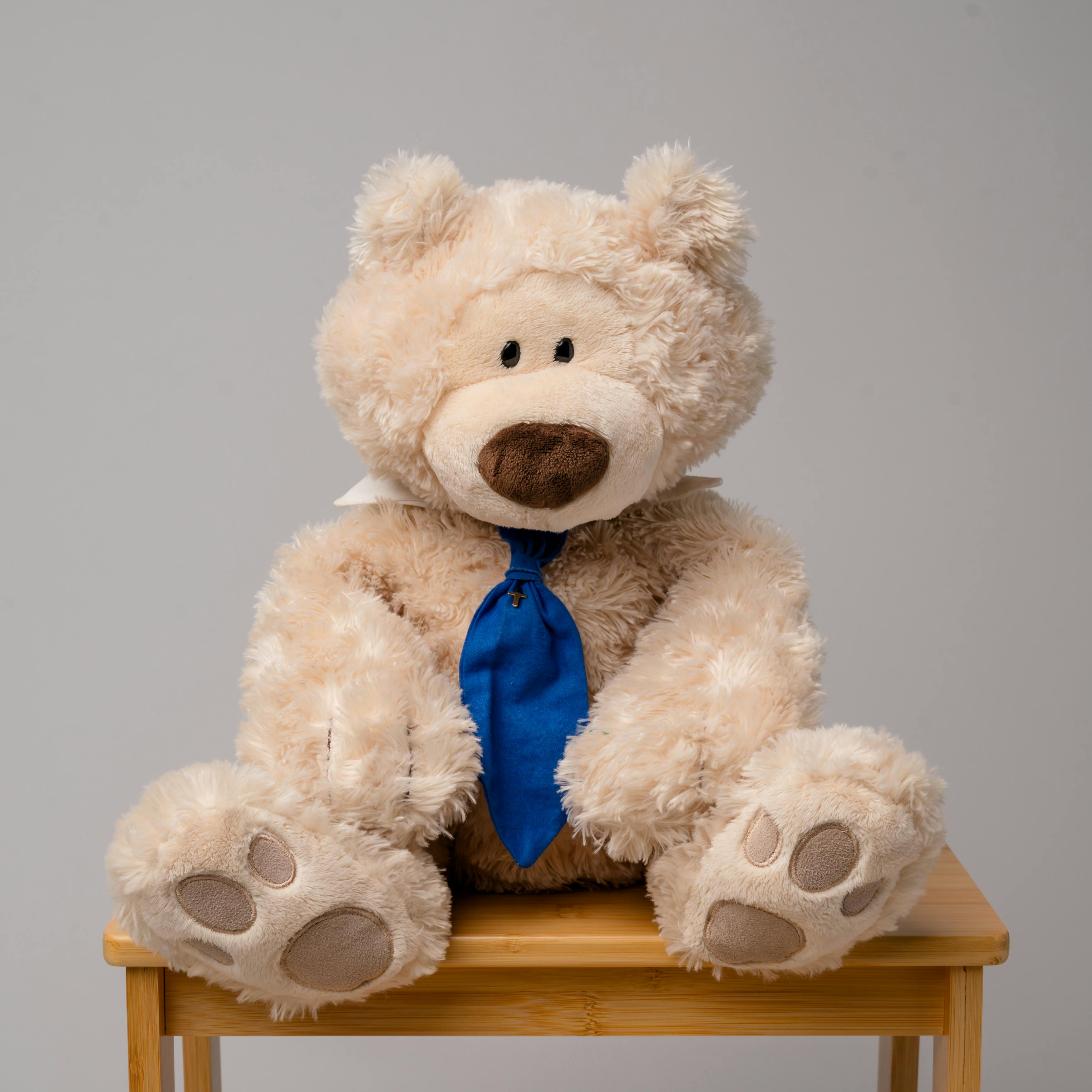 Tan teddy bear wearing a white collar and blue tie, sitting on a wooden stool