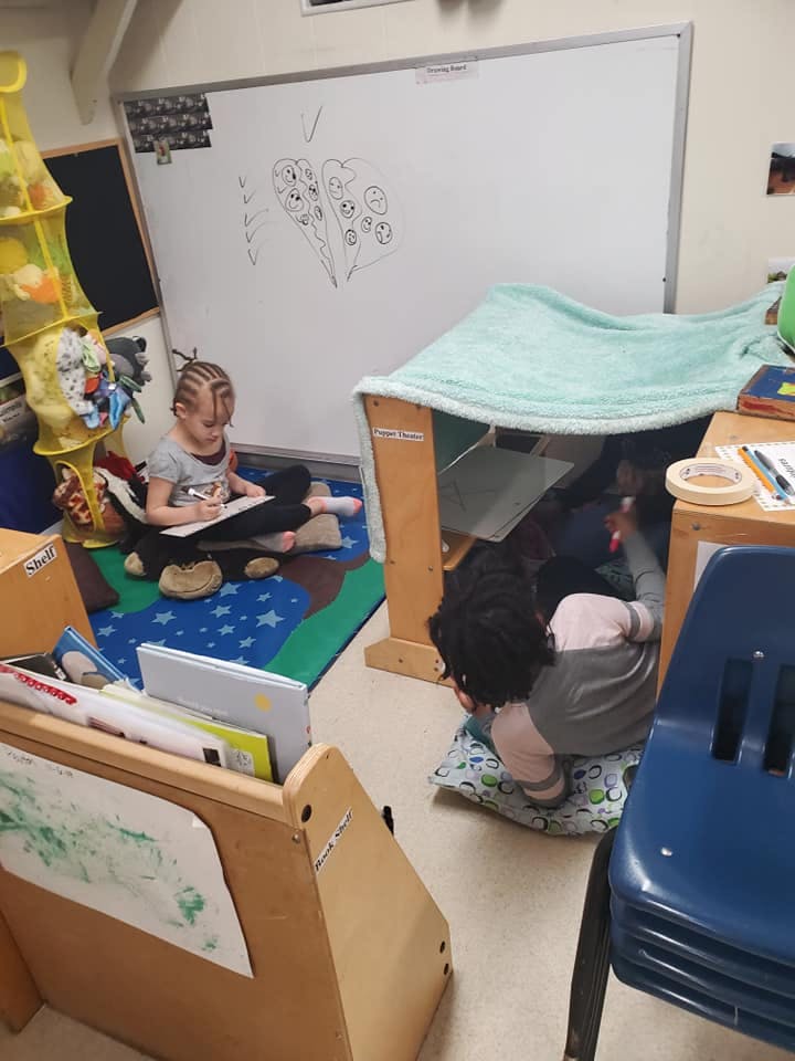 Two children playing in a classroom, one drawing, one in a fort