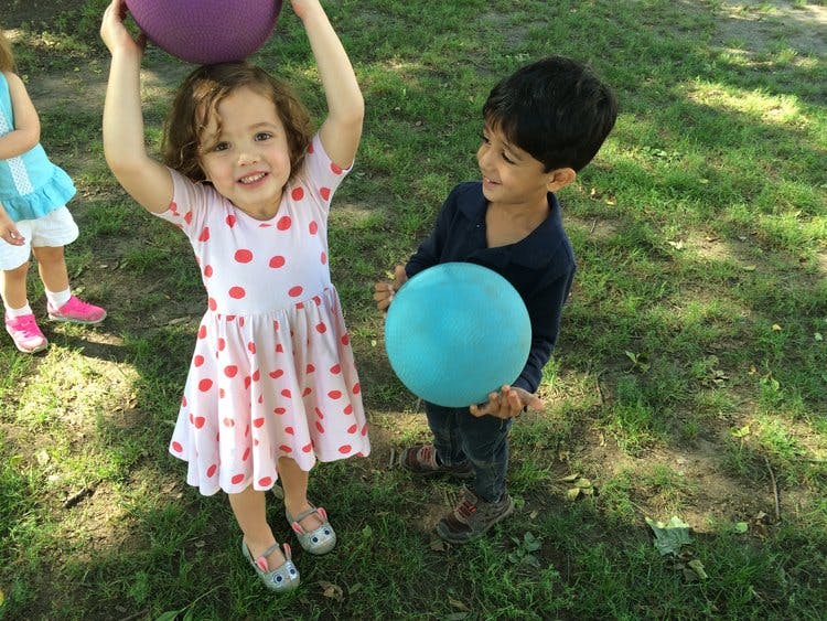 Child holding a purple ball overhead, another child holding a blue ball