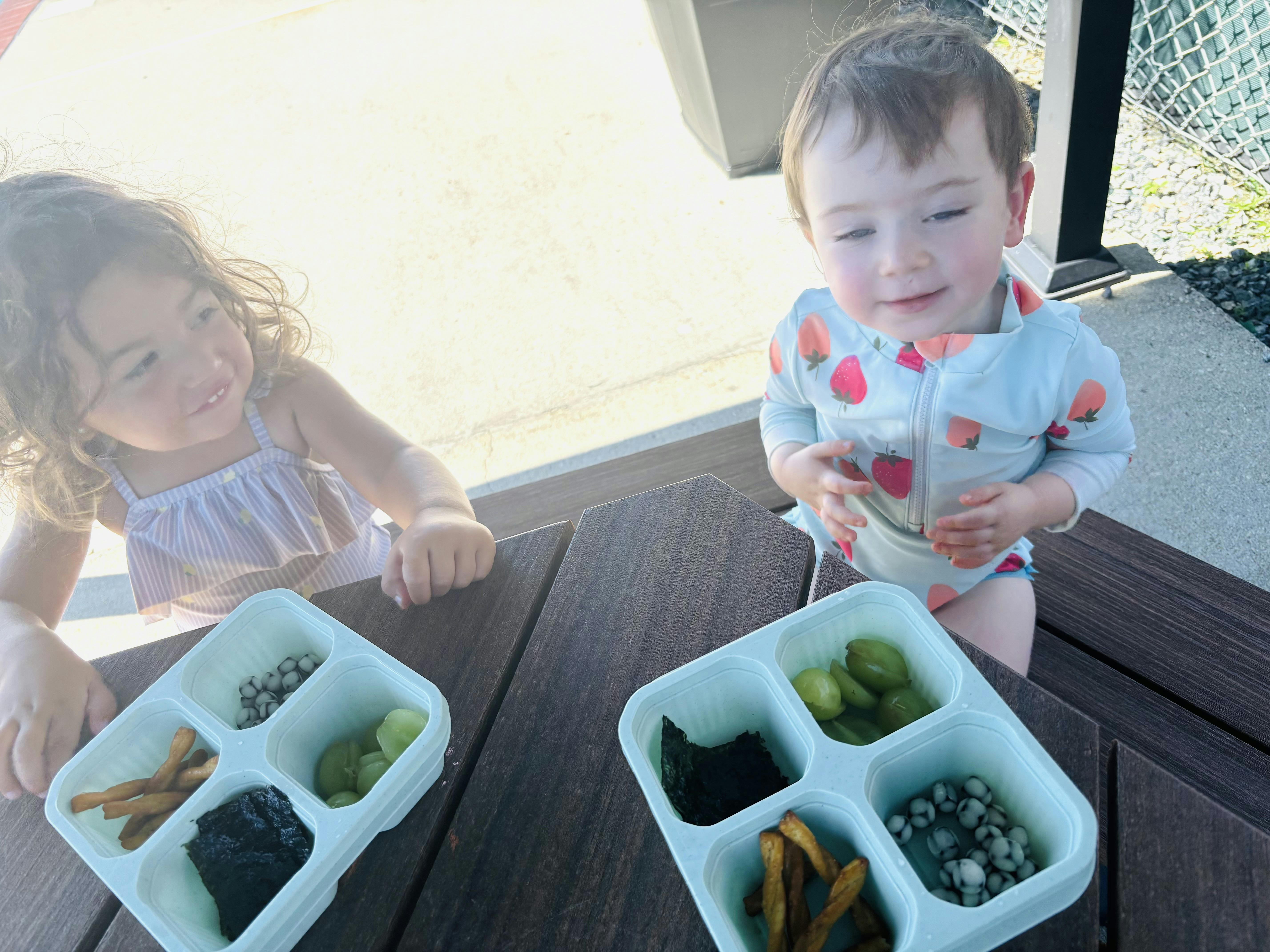 Two children sitting at a picnic table with trays of snack food