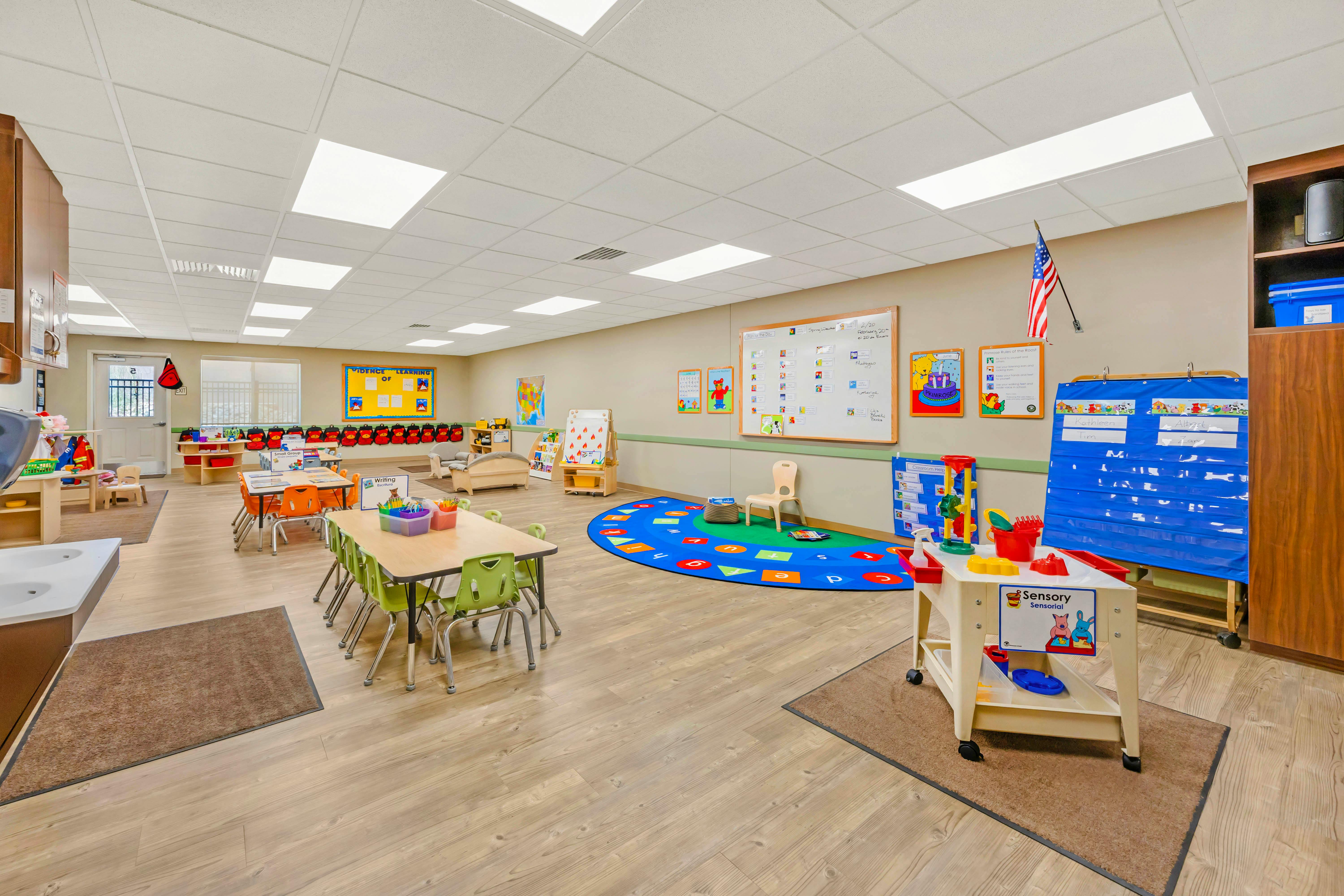 Empty classroom with small tables and chairs, a play area rug, and learning materials