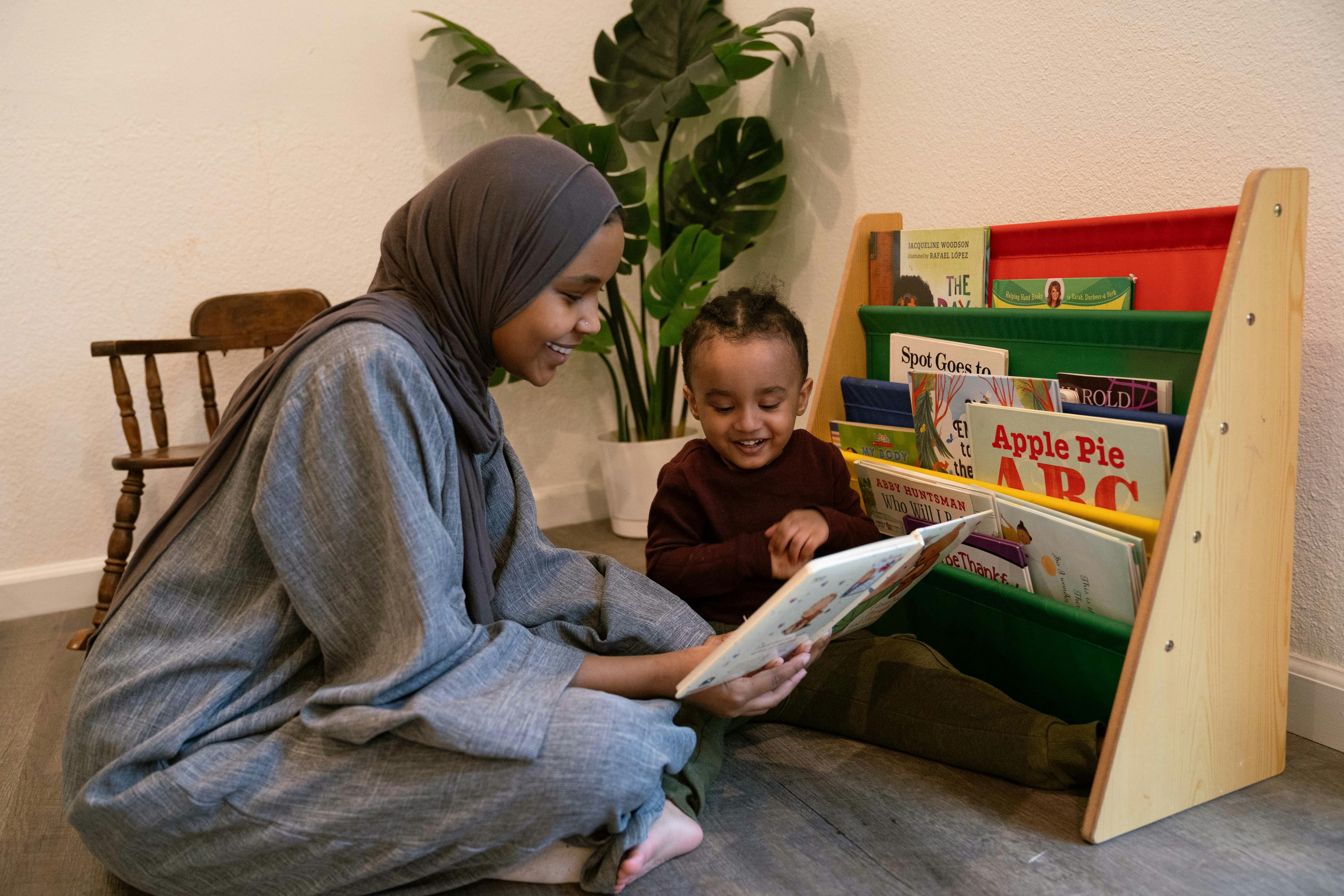 Person in hijab reading a book to a child next to a bookshelf