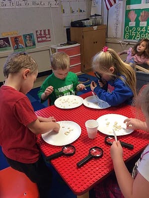 Children at a red table with plates of small objects and magnifying glasses