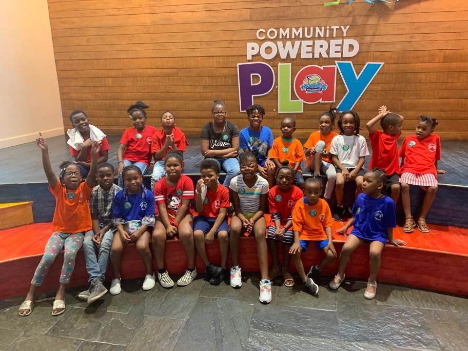 Group of children sitting and standing in front of a "Community Powered Play" sign