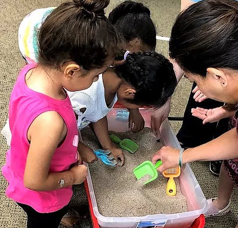 Children and an adult playing with sand and shovels in a container