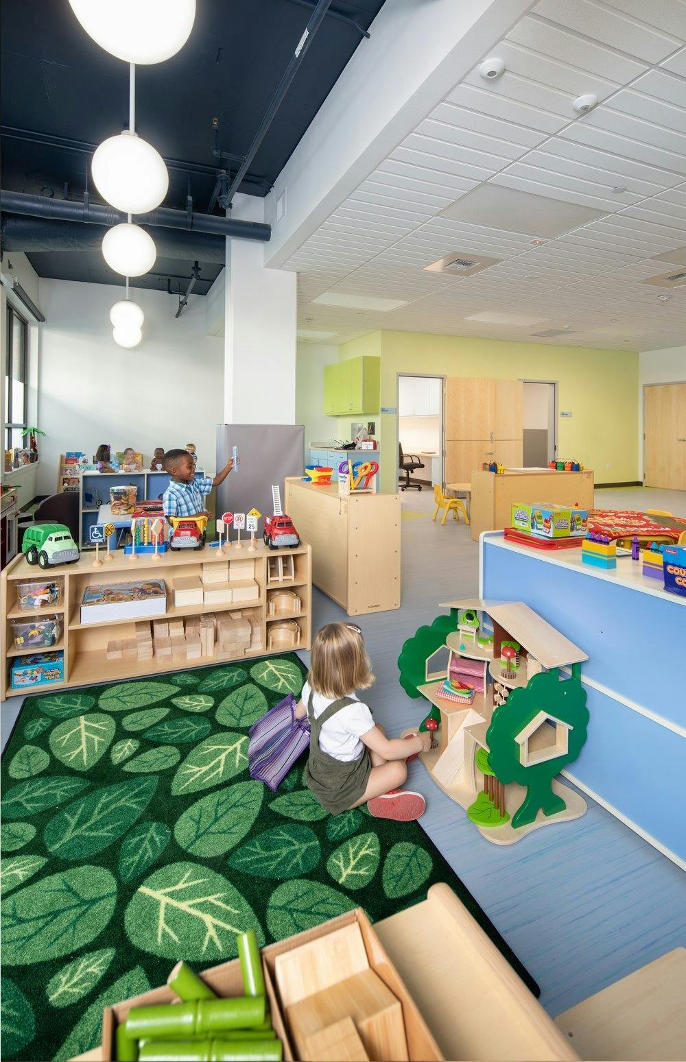 Child playing with a treehouse dollhouse on a leaf-patterned rug in a classroom