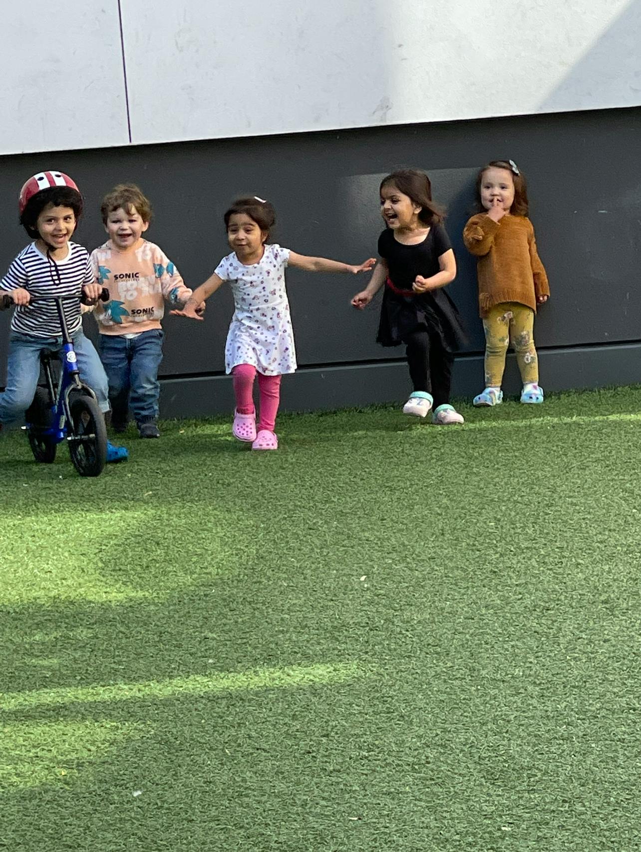 Five children playing on artificial grass, one on a small bicycle