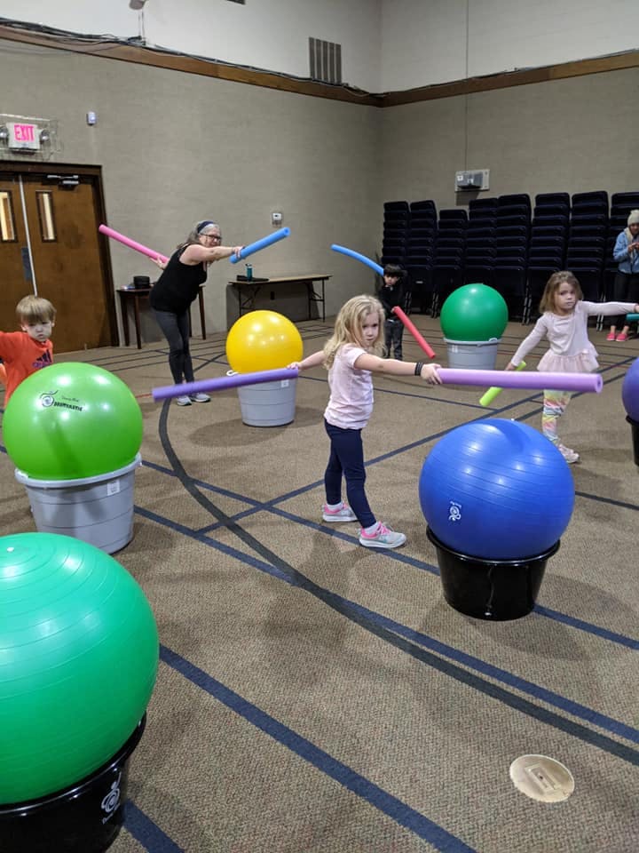 Children and an adult holding foam noodles in a room with exercise balls