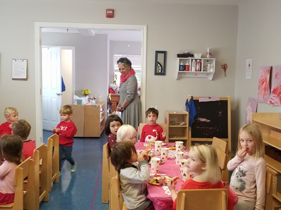 Children sitting at tables in a classroom with an adult standing in a doorway