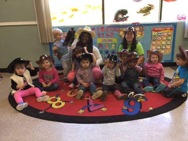 Two adults and several children sitting on a rug, some wearing hats