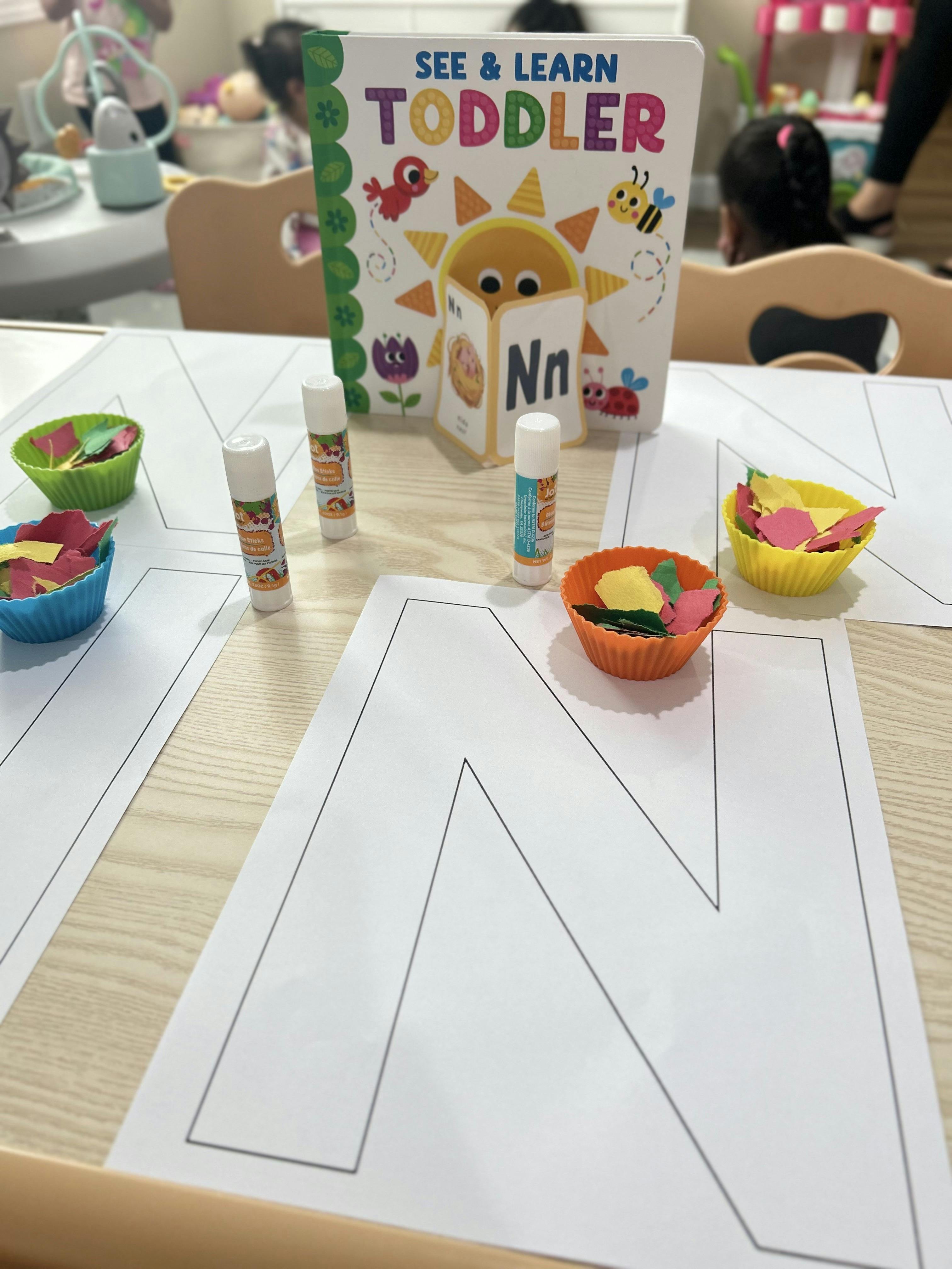 Craft supplies on a table including a toddler book, glue sticks, and paper cut-outs