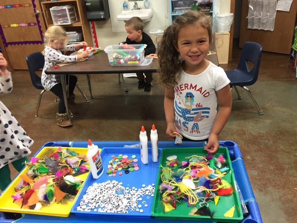 Child standing at a craft table with supplies, "Mermaid" on shirt
