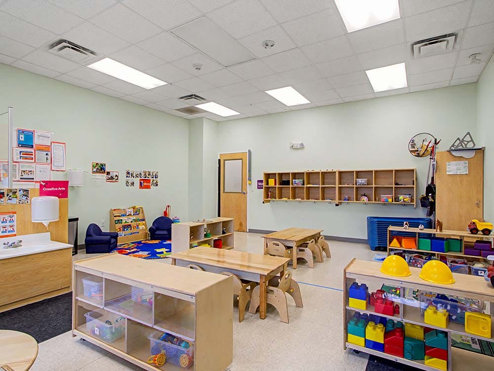 Empty classroom with small tables and chairs, shelves with toys, and cubbies