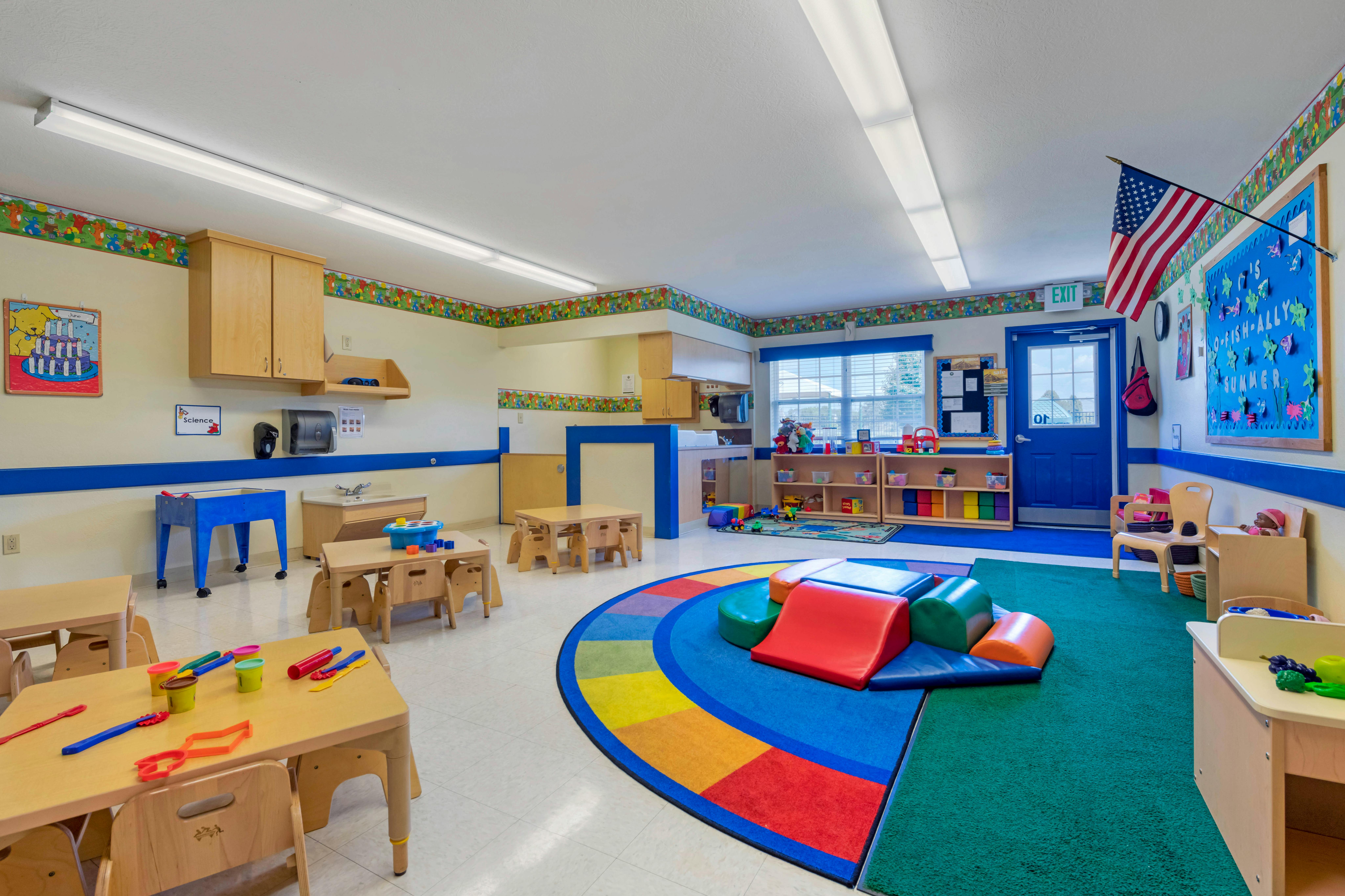 A brightly lit classroom with small tables and chairs, a play area, and storage shelves