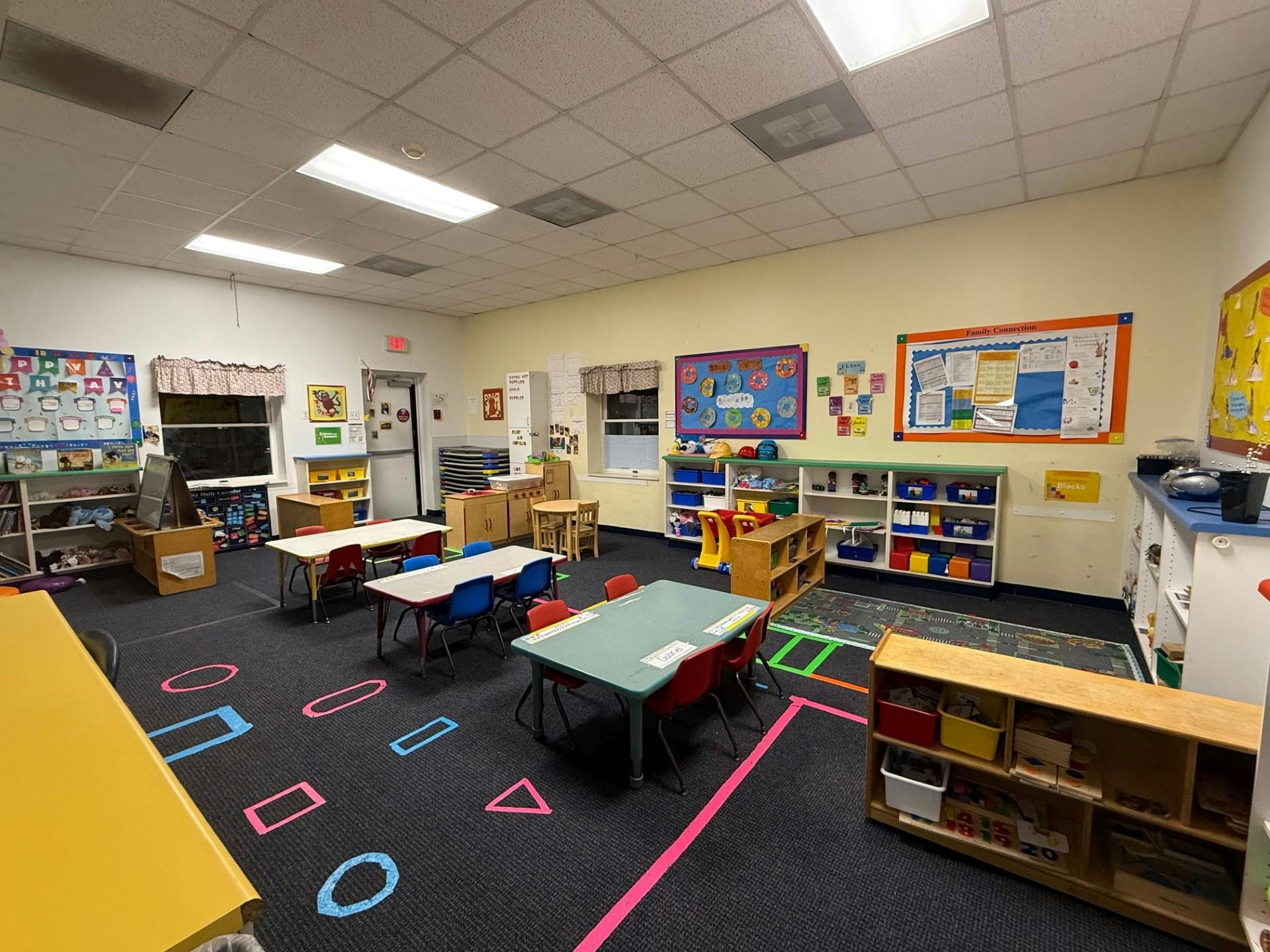 Classroom with small tables and chairs, shelves with toys, and colorful shapes on the floor