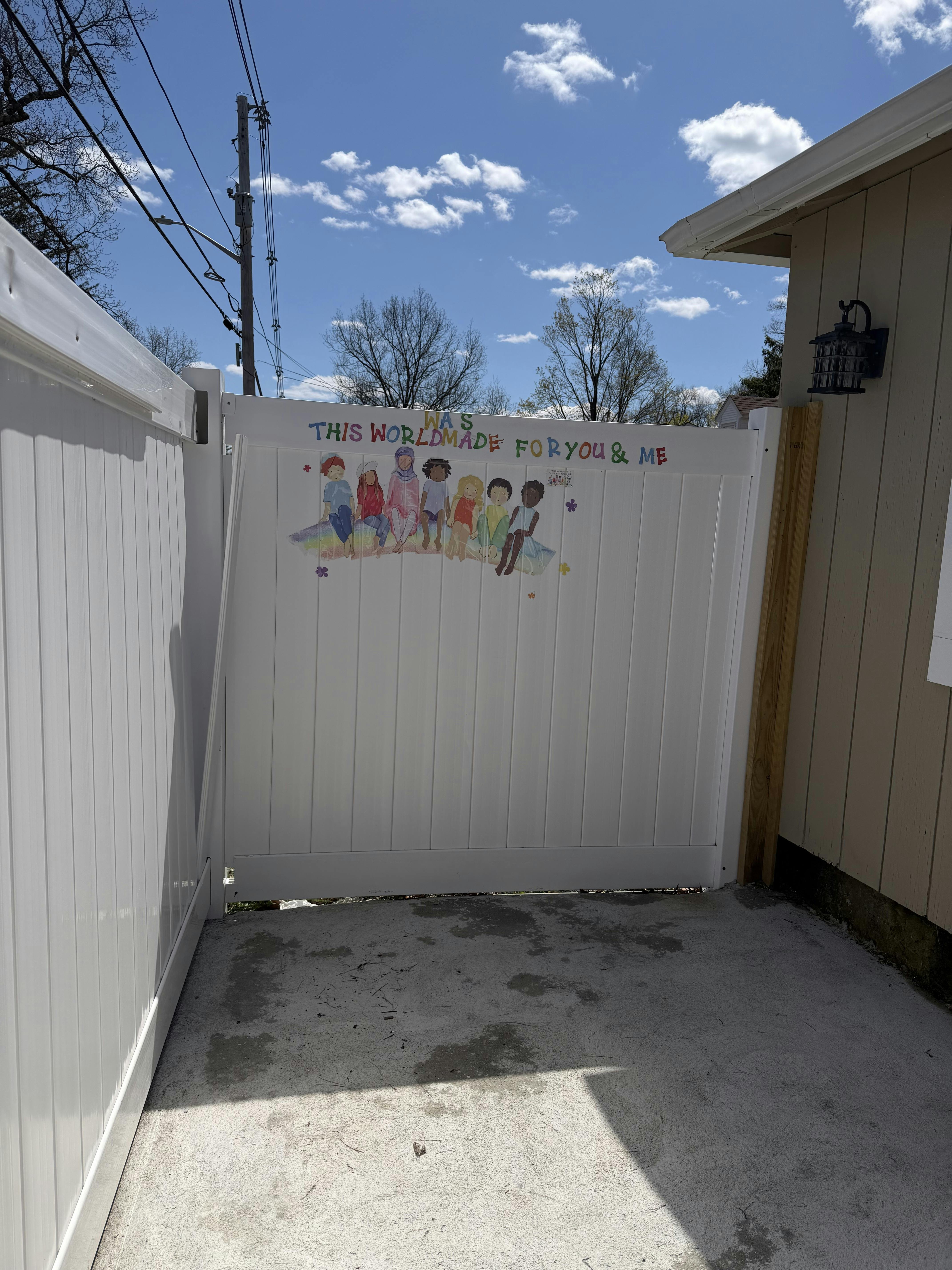 White fence gate with a colorful drawing of children and text that says THIS WORLD MADE FOR YOU & ME