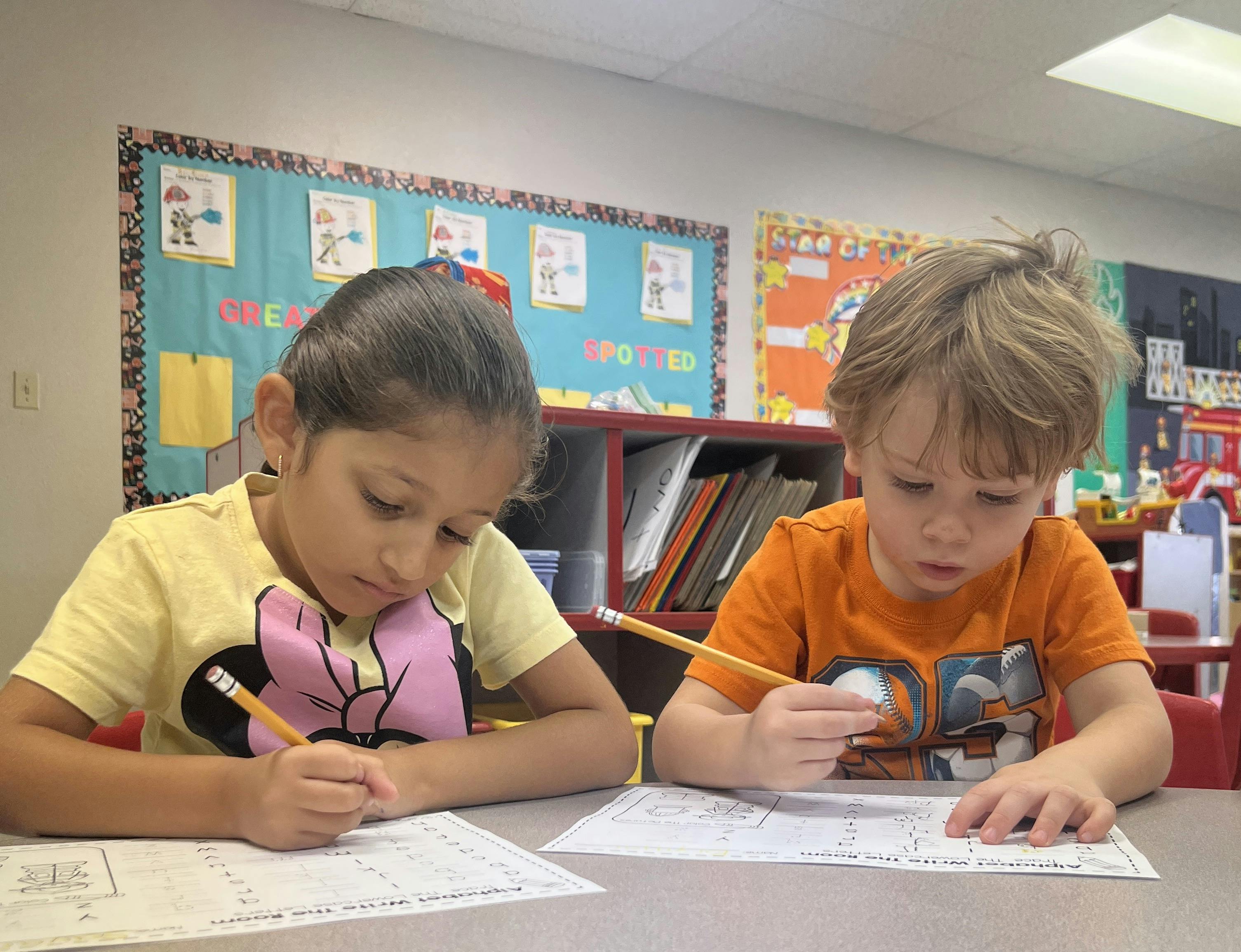 Two children writing on papers with pencils in a classroom